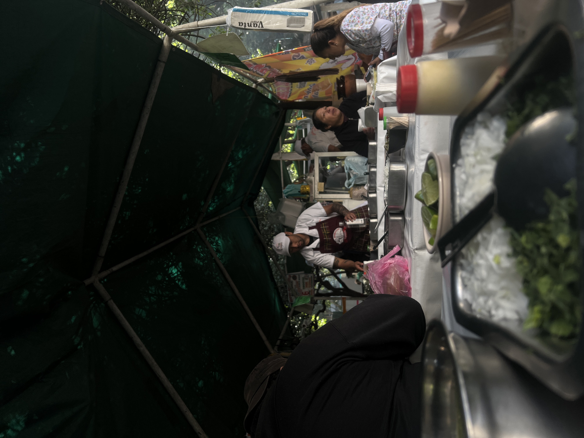 The image shows a food stand in Mexico City, Mexico. The stand is under a green canvas tarp, and several people are present. A man wearing a white chef's hat and a red plaid apron is working at the stand. Two women appear to be working together, while another woman seems to be waiting for service. There are various containers and utensils on the stand, including containers of limes, chopped onions, and chopped herbs. Condiment bottles are also visible. A person in a black hooded sweatshirt stands in the foreground, partially obscuring the view of the stand. A box of "Vanta" paper towels hangs from the tarp.