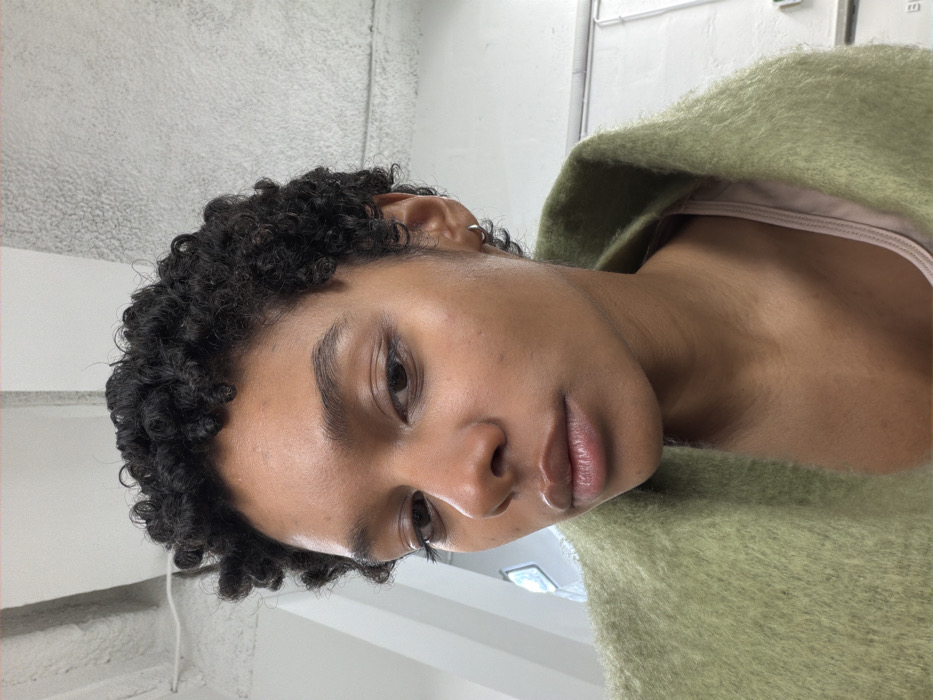 A close-up portrait features a female individual with dark skin, short, tightly coiled black hair, and dark eyes, gazing directly forward. She wears a light olive-green, fuzzy outer garment, appearing to be a jacket or coat, over a light pink or nude-colored inner garment. A small, thin silver hoop earring is visible in her right earlobe. The background is an indoor setting with a light-colored, possibly concrete, ceiling displaying an exposed white pipe or conduit in the upper left. The wall behind her is textured and light grey. A blurred light source or window is partially visible on the far left, and a white, flat panel or door is present on the right. No discernible landmarks or specific features are present within the image to identify a location within Paris, France.
