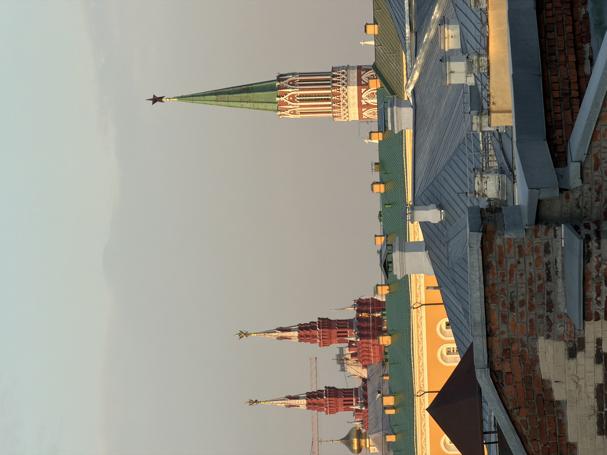 This image shows a rooftop view of Moscow, Russia, with several iconic Kremlin towers visible in the background. The foreground features a brick wall and various rooftops, some with metal cladding and chimneys. In the mid-ground, there are buildings with green and yellow roofs. Dominating the background are the red towers of the Moscow Kremlin, topped with golden eagles, and a tall, green-spired tower crowned with a red star. The sky is a pale, hazy blue, suggesting either early morning or late afternoon light. There are no people visible in the frame.