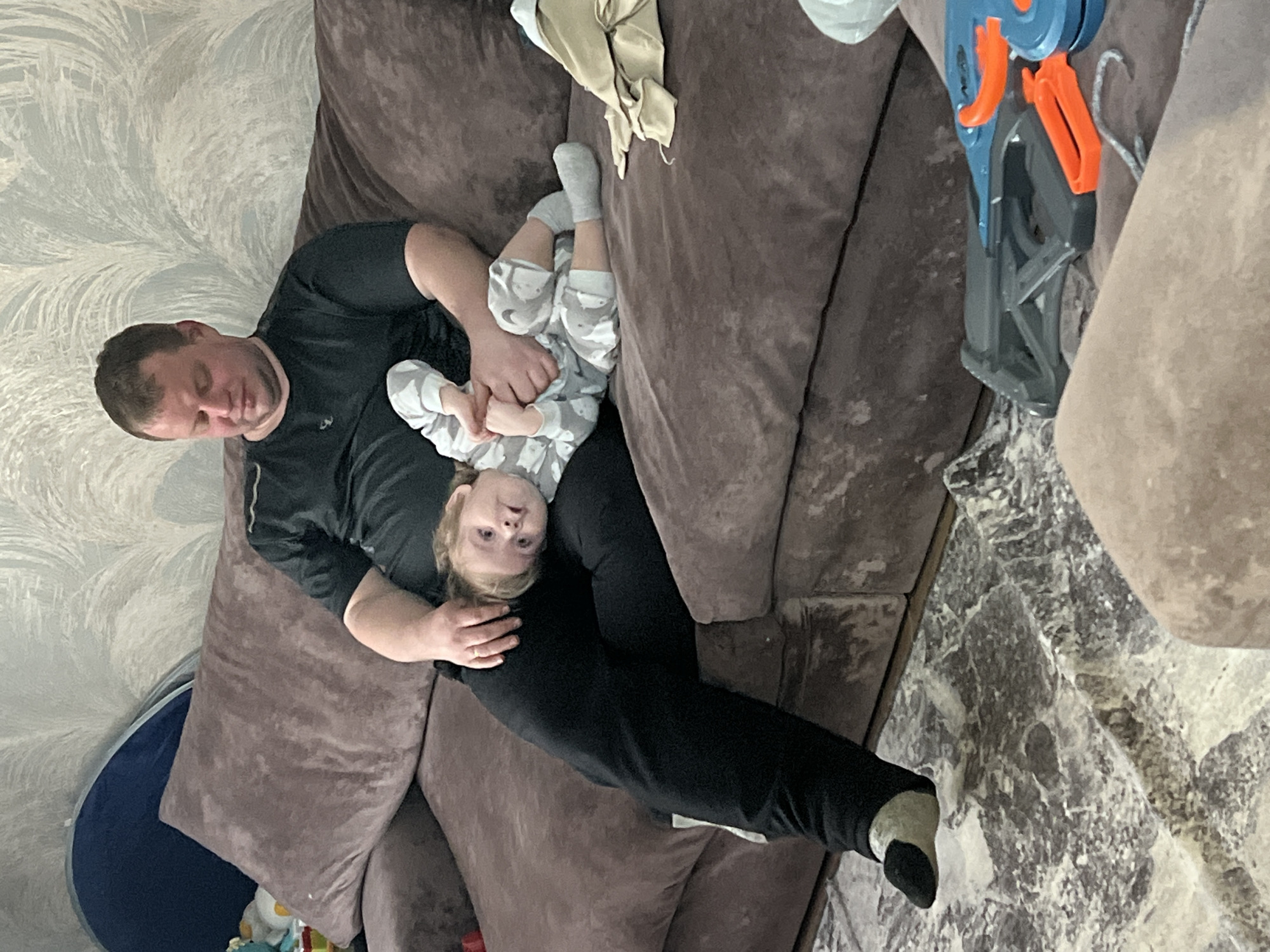 An indoor scene in a residential setting in Noviy, Russia, depicts a male adult and a young child on a brown upholstered sofa. The adult male, with short brown hair and wearing a dark t-shirt and black sweatpants, is seated on the left side of the sofa, looking downwards with his eyes closed or partially closed. He wears a ring on his left ring finger. His left hand rests on the child's head, and his right hand holds the child's feet. The child, a toddler with light-colored hair and wearing a grey and white patterned romper and grey socks, is lying on their left side across the man's lap and the sofa, looking directly at the camera with wide eyes. The child's hands are clasped near their chest. Behind the sofa, the wall is covered with light-toned wallpaper featuring a grey and white feather-like pattern. To the far left, a dark blue fabric structure is partially visible behind a sofa cushion. On the right side of the sofa, a crumpled piece of beige fabric is present. The floor in the foreground is covered by a grey and white patterned rug, and a blue and orange toy blaster, possibly a Nerf gun, along with a grey plastic toy component, are visible on the floor to the lower right. The scene conveys a relaxed, domestic environment.