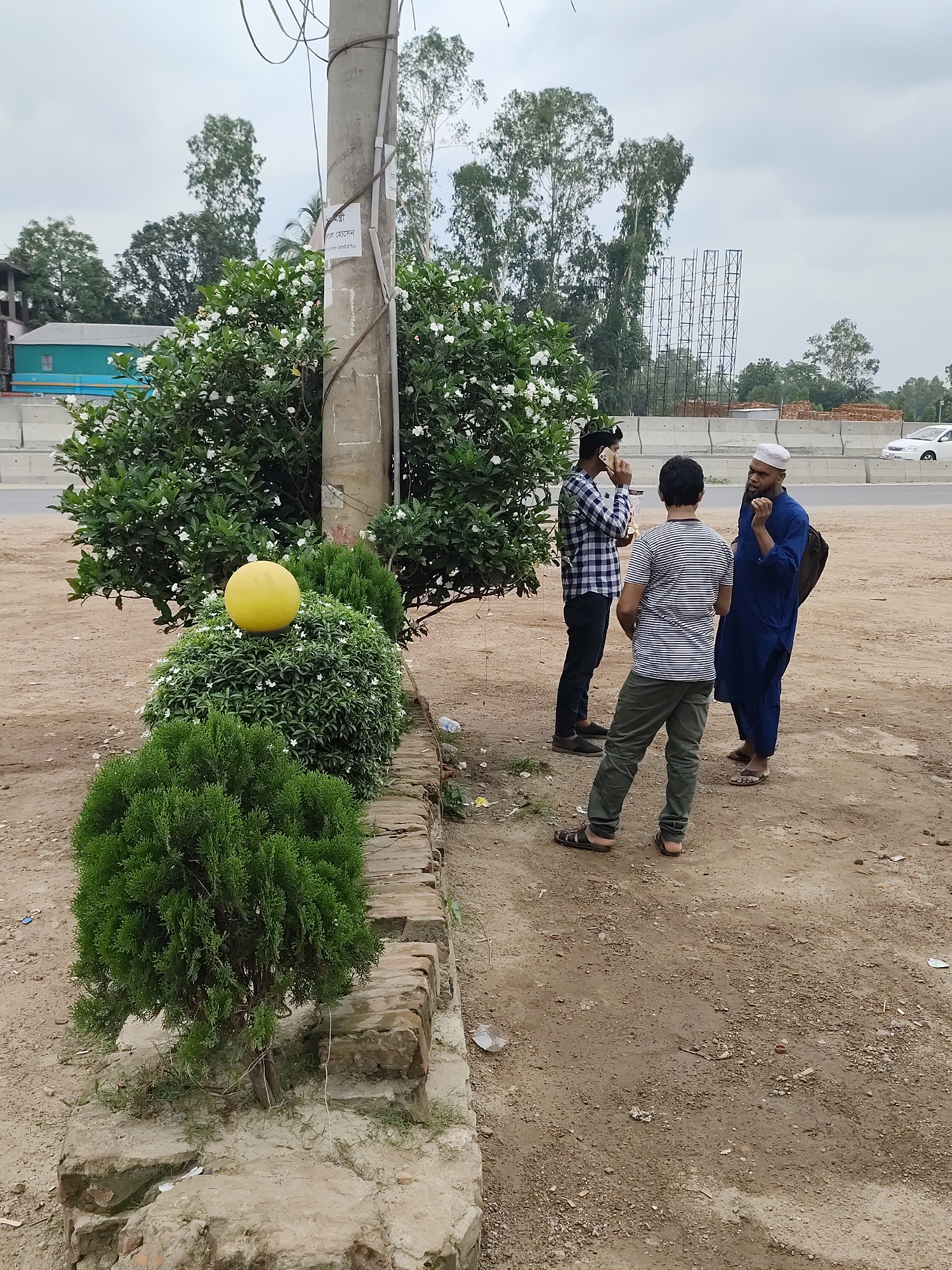 The image shows three men standing on a dirt patch in Hatikumrul, Bangladesh. The man on the left is on his phone, wearing a checkered shirt and black pants. The man in the middle wears a striped shirt and green pants. The man on the right is wearing a blue robe and a white cap. They are standing next to a small brick wall and a bush with white flowers. A yellow ball sits atop the bush. A utility pole is behind the bush, with wires running from it. Behind the men, there is a road with a concrete barrier and some vehicles. Trees and buildings are visible in the background.