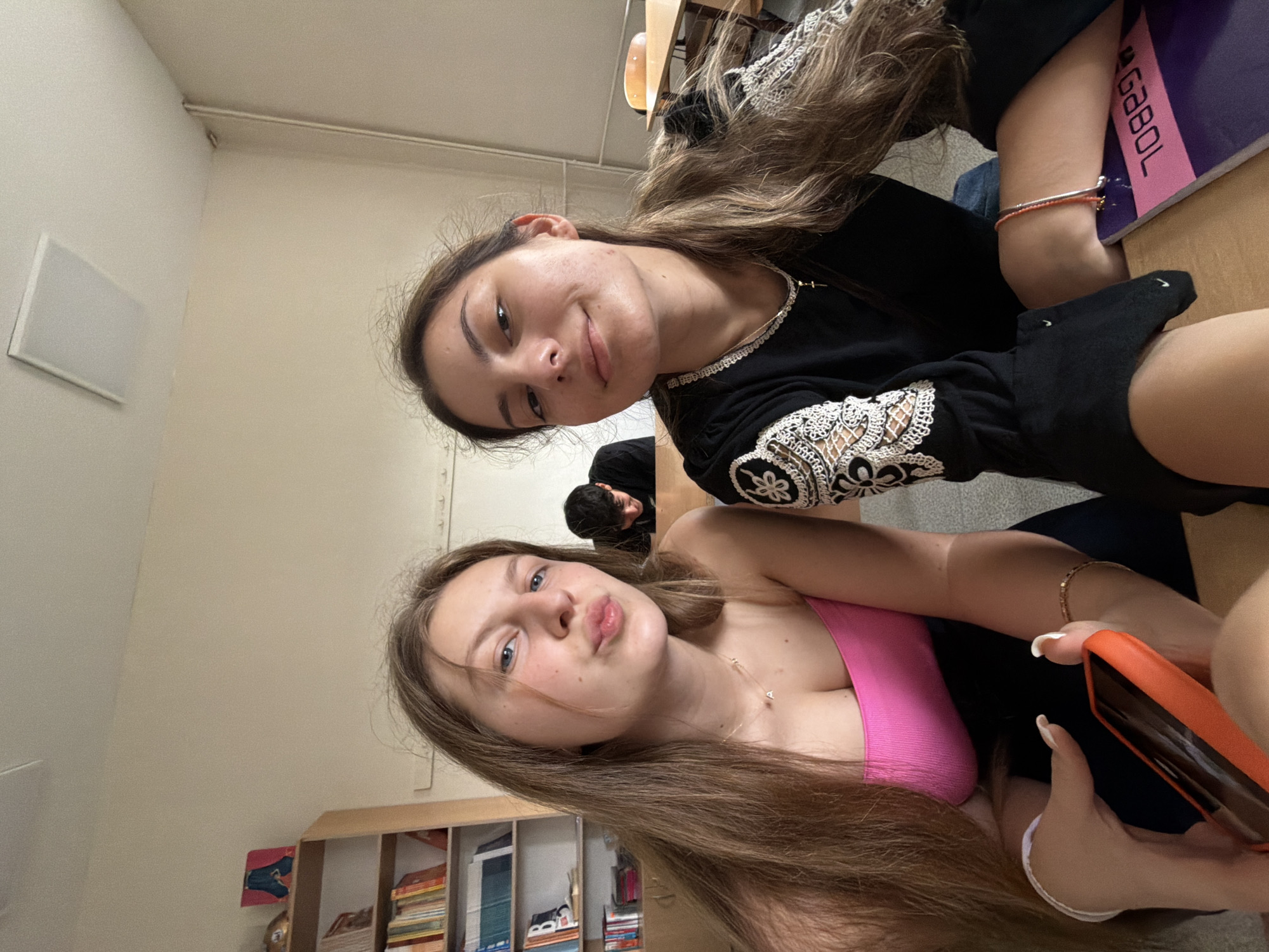 This indoor photograph, taken in Burgas, Bulgaria, captures two young women posing for the camera, likely within a classroom or study setting. The image is oriented with the subjects tilted.
In the foreground, a young woman with long, light brown hair and blue eyes looks directly at the viewer with puckered lips, making a playful "kissing face." She is wearing a bright pink sleeveless top and a delicate necklace. Her right hand, featuring white-tipped nails, holds an orange smartphone, suggesting she is taking the photo.
Beside her, another young woman with long, dark brown hair smiles subtly. She is dressed in a black long-sleeved top adorned with intricate white lace embroidery on the sleeves and neckline. She wears a thin necklace with a small cross pendant and two thin bracelets (one orange, one dark) on her left arm. Black socks with a white Nike swoosh are visible on her feet.
In the mid-ground, a third person with dark hair is partially visible from behind, seated at a desk. The background elements consistent with a learning environment include light-colored walls, wooden desks, and bookshelves stocked with various books. A white rectangular panel, possibly a speaker or ventilation cover, is mounted on the wall.
A notable object is a purple notebook or binder resting on a desk, clearly displaying the brand name "GABOL" and the number "70899" below it. The overall mood conveyed by the subjects' relaxed expressions is friendly and informal.