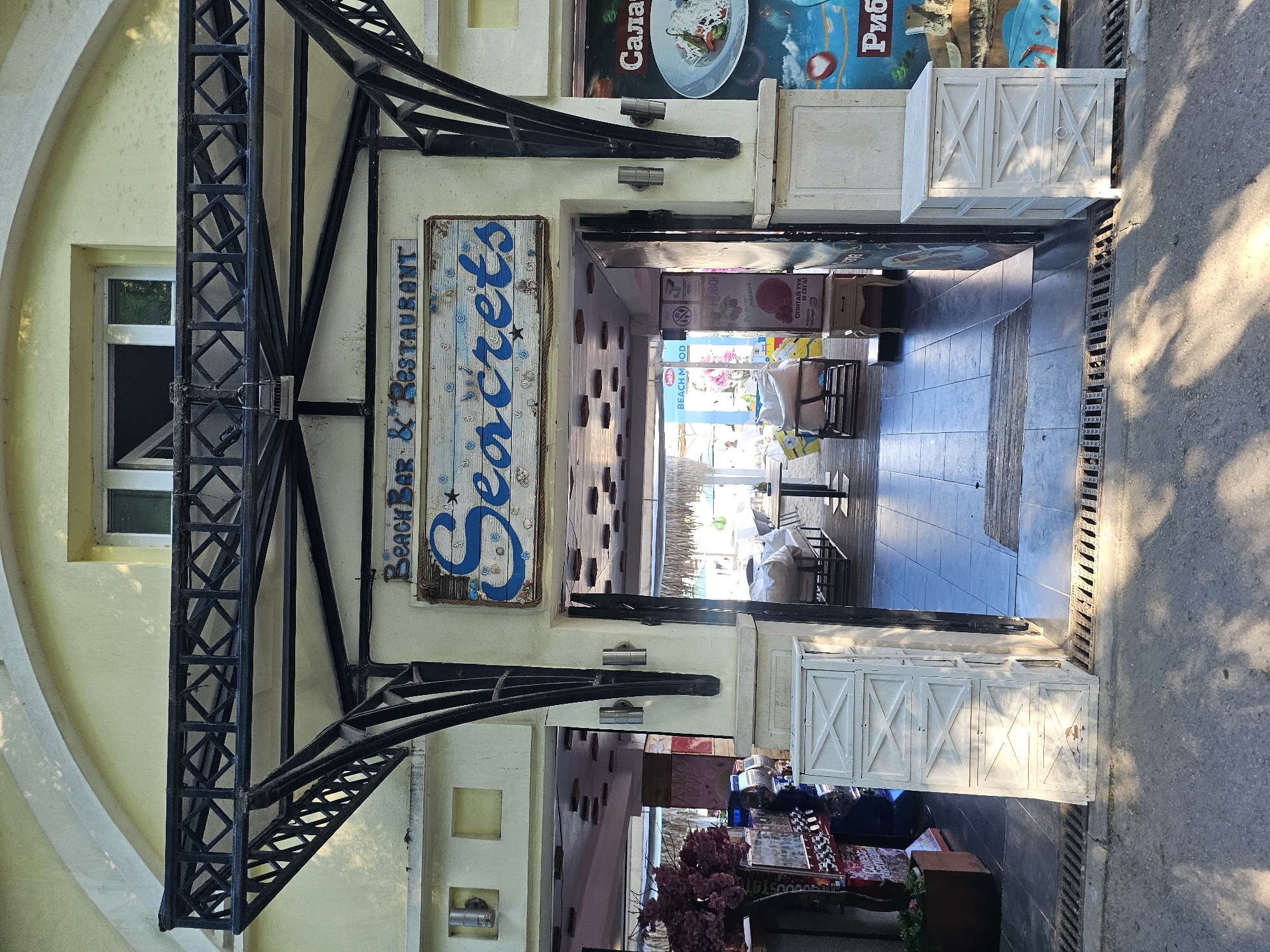 The image shows the entrance to a beach bar and restaurant named "Seacrets". The facade is painted a pale yellow with a large arched entryway. Above the entrance, a black wrought-iron structure supports an awning. A sign with blue lettering reads "Beach Bar & Restaurant" and below it, a larger sign features the name "Seacrets" in blue script on a distressed wooden background adorned with shells and nautical motifs. Through the open doorway, tables and chairs are visible within a covered outdoor seating area. To the right of the entrance, a colorful mural depicts food items and text in an unfamiliar language. Two white, decorative cabinets with criss-cross paneling are positioned on either side of the entrance. The ground in front of the establishment is paved with dark tiles and a metal grate runs along the edge of the walkway. Shadows of trees are cast on the ground and the building.