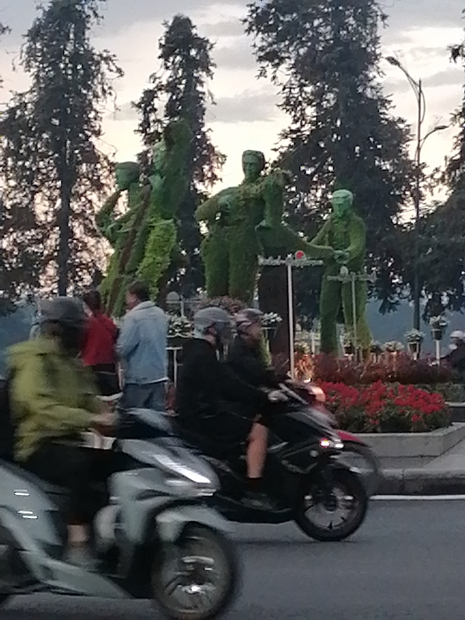 The image shows a street scene in Da Lat, Vietnam. In the background are topiary sculptures of people, arranged in a park-like setting with flower beds. Taller trees loom behind the sculptures. In the foreground, two motorbikes are moving along the street. The riders are wearing helmets and casual clothing. Other people are visible in the background near the sculptures. The overall lighting suggests it is dusk or early morning.