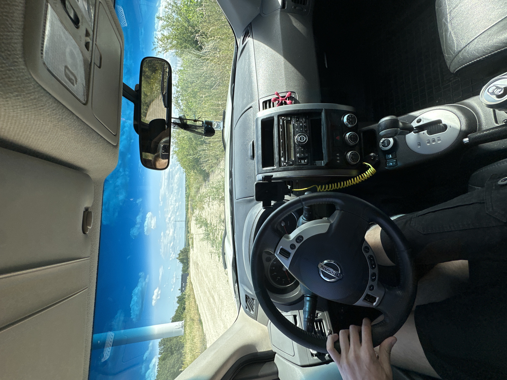 The image is a first-person perspective from inside a car, looking forward. The driver's hands are visible on the steering wheel, which has a Nissan logo. The dashboard features a radio, climate controls, and a gear shift. A rearview mirror reflects the road ahead, which appears to be a dirt track in a rural area with greenery and a blue sky with clouds. In the distance, a large structure, possibly a wind turbine, is visible on the left. The car is likely located on the outskirts of Abrykosivka, Ukraine, given the presence of rural landscape features.