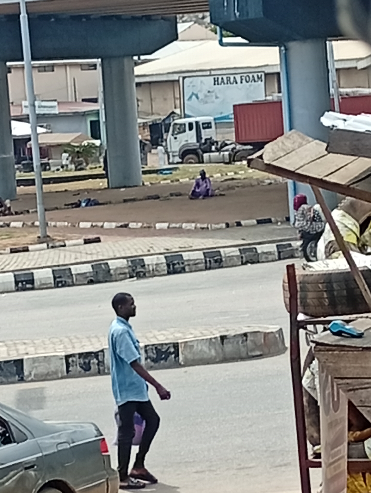 The scene depicts a city street. A man in a light blue shirt and dark pants walks along the side of the road, near a parked gray car. In the background, under an overpass structure, a truck is parked near a building labeled "HARA FOAM." A woman in a purple outfit is sitting on the ground. Other people are visible in the background. The environment seems urban, with other buildings and infrastructure elements visible in the distance.
