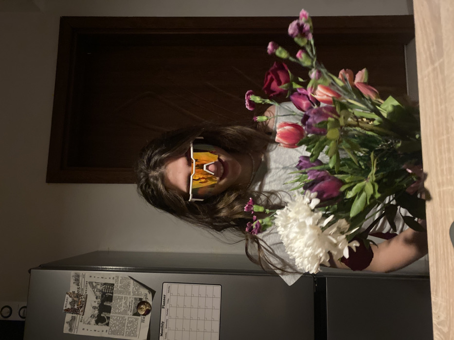 The image depicts an indoor, dimly lit scene. A young individual, with long brown hair, occupies the central frame. They are wearing a grey t-shirt and a pearl necklace. White-framed, mirrored sports sunglasses cover their eyes, reflecting an indistinct interior, while a subtle smile is visible on their face. The individual holds a substantial bouquet of various flowers, prominently featuring white chrysanthemums, purple and pink tulips, red roses, and smaller pink/purple carnations. In the left background, a silver-toned refrigerator is visible, adorned with a newspaper clipping that includes text fragments such as "MILANO" and "Bruxelles," and a magnetic "Monthly Planner" calendar. To the right, a dark brown wooden door is closed within its frame. A light-colored wooden surface runs across the immediate foreground. The scene is located in an interior room in Sofia, Bulgaria.