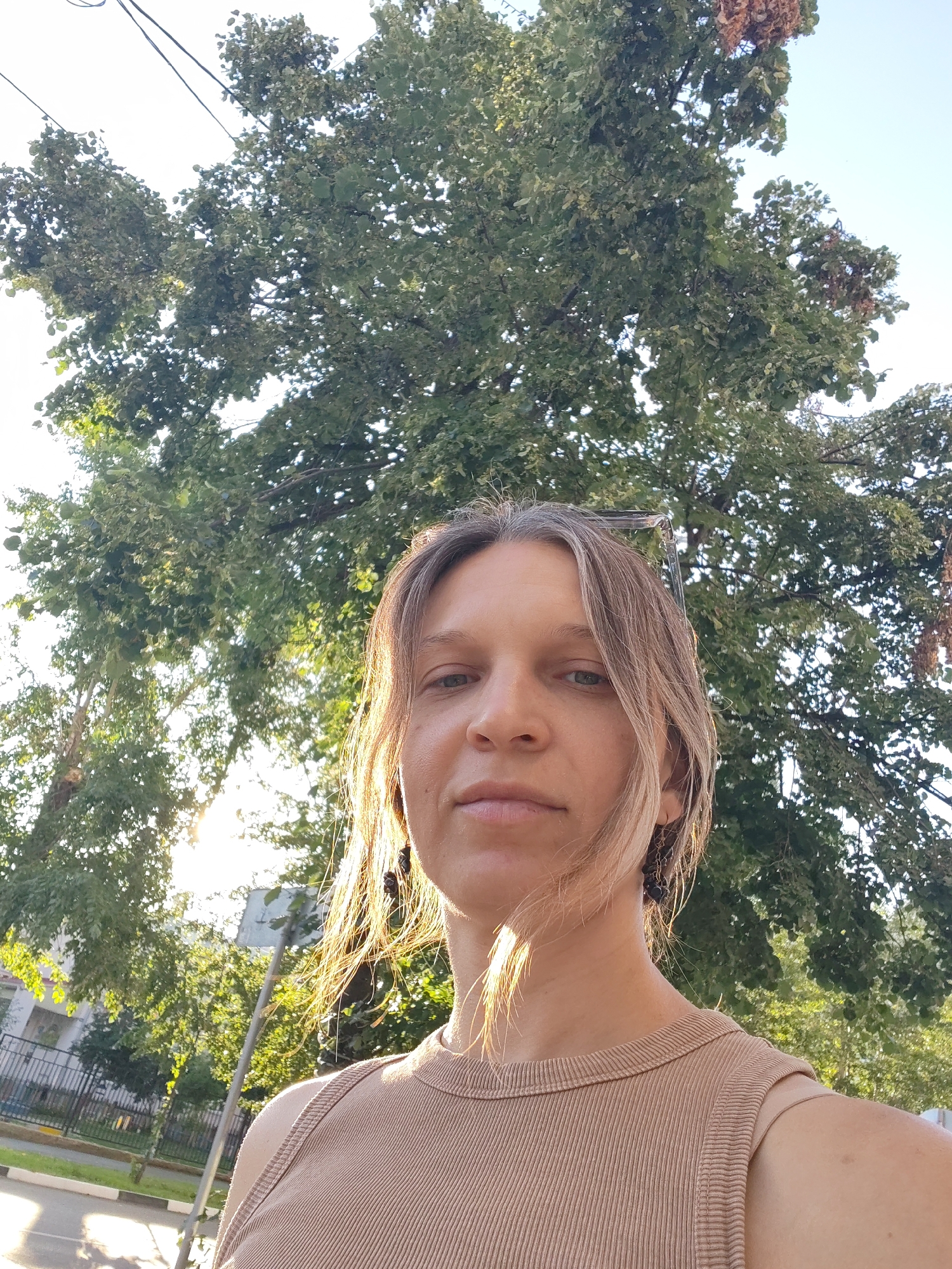 A medium shot captures a woman from the shoulders up, taken outdoors in what appears to be an urban setting, possibly a park or street with trees. She is looking towards the camera with a neutral expression. The lighting suggests it is daytime, with sunlight filtering through the leaves of a large tree behind her, casting a warm glow on her face and hair. Her hair is light brown and styled loosely, with some strands falling forward. She is wearing a ribbed, sleeveless top in a neutral tan color. In the background, out of focus, are green trees, some buildings, a fence, and what looks like a street with a sidewalk. The overall impression is a candid shot taken in natural light.