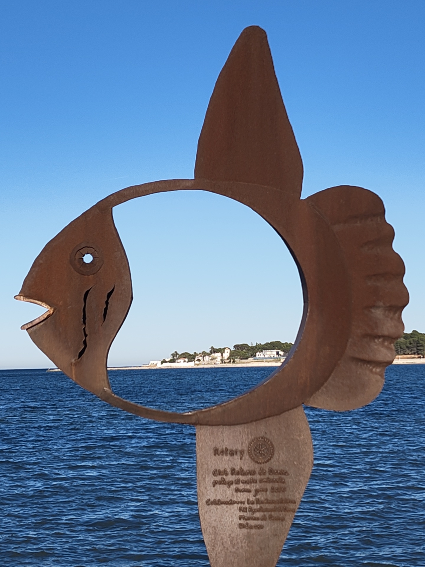 The image shows a rusty, metal sculpture of a fish with a large hole in the center, positioned near the coast. The fish is mounted on a short pedestal, which has the "Rotary" logo and some inscriptions in Spanish. Through the hole in the fish, a stretch of land can be seen with white buildings and trees. The sculpture is set against a background of blue sea and a clear blue sky. The location is likely Dénia, Spain, as the sculpture is located there.