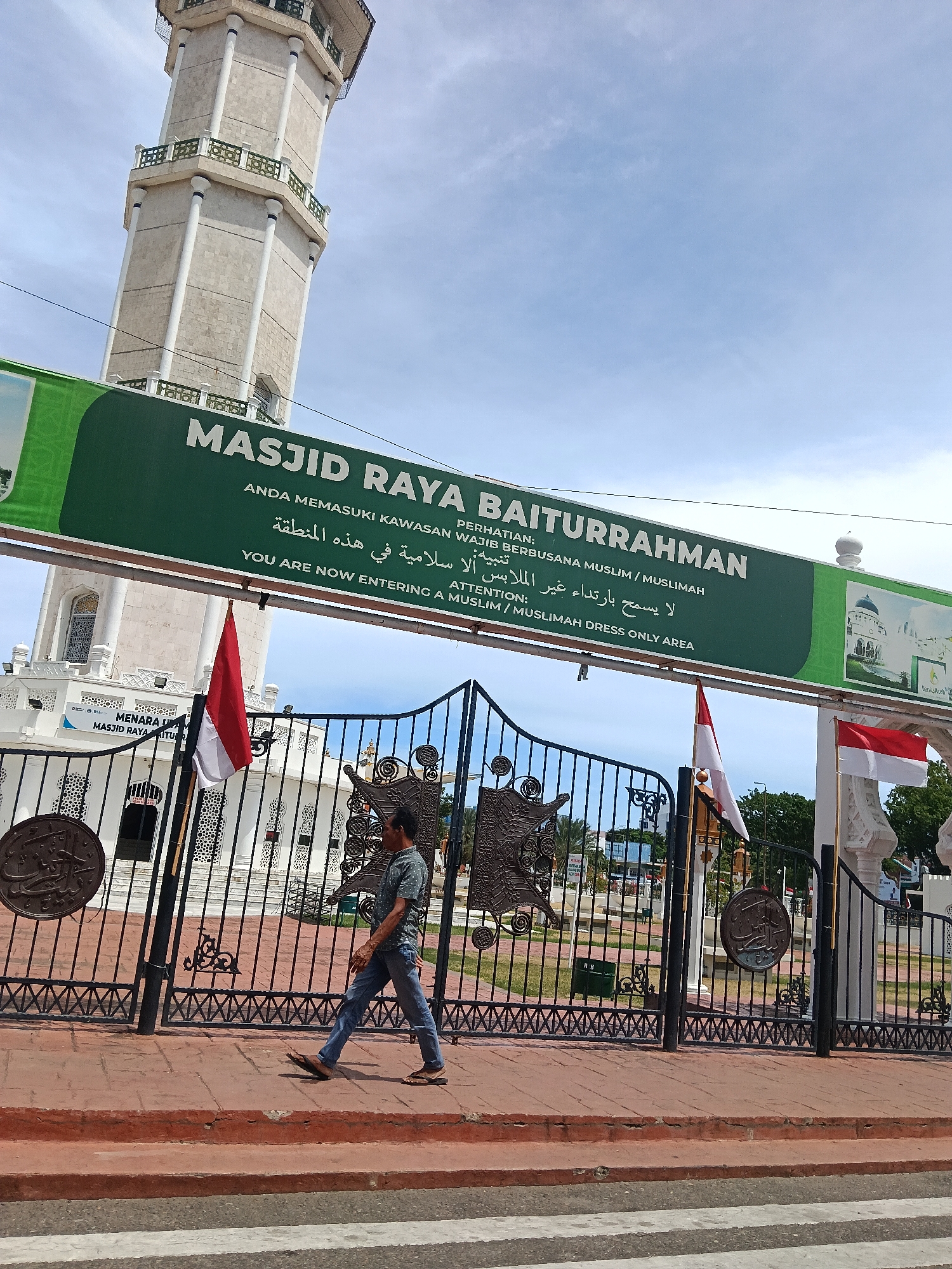The image depicts the entrance to Masjid Raya Baiturrahman in Banda Aceh, Indonesia. A man in jeans and a patterned shirt is walking in front of a black metal gate. The gate has decorative elements and is adorned with two flags, one Indonesian flag and one with red and white stripes. Behind the gate, a white building with intricate architecture and a tall, slender minaret can be seen. A large green banner with text in Indonesian, Arabic, and English is prominently displayed above the gate, indicating that this is a Muslim/Muslimah dress-only area. The sky is clear with a few wispy clouds. The ground in front of the gate is paved with reddish bricks and a crosswalk is visible at the bottom of the image.