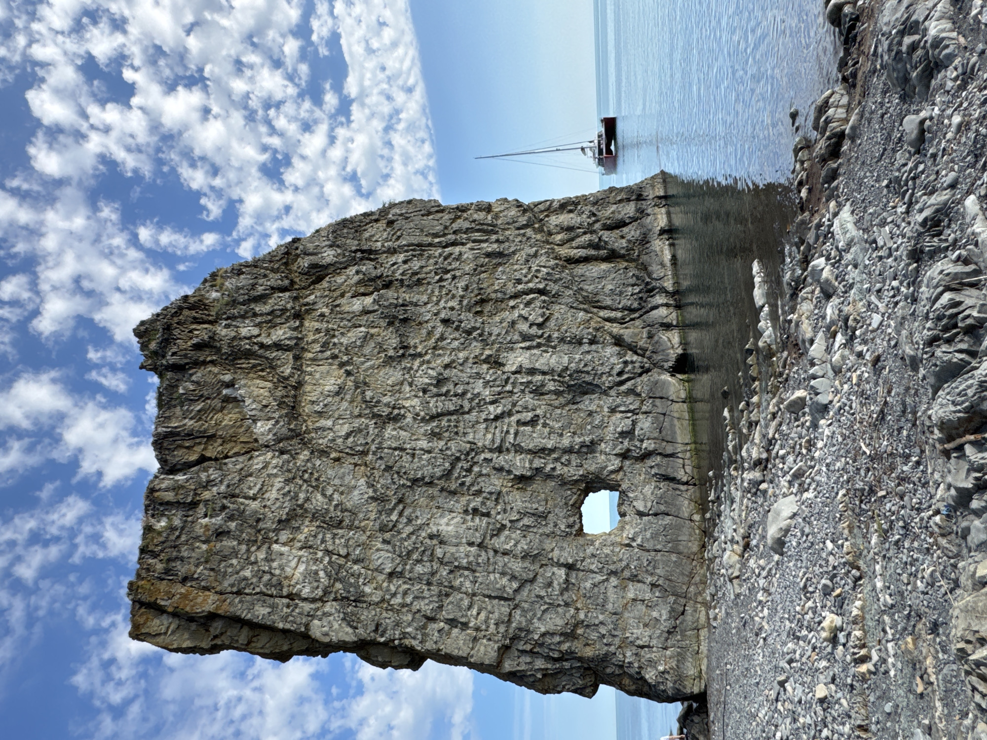 The image, when correctly oriented, captures a serene coastal landscape under a bright, clear sky. In the foreground, a massive, rugged cliff or rock formation dominates the left and central parts of the frame. Its surface is heavily textured with numerous layers and cracks, appearing in shades of grey and earthy brown. A distinctive natural opening, roughly rectangular or square, is visible within the rock towards its right side, offering a glimpse of the blue sky and distant sea beyond.
To the right of the rock formation, the calm, blue-grey sea extends into the horizon, reflecting the bright sky. A solitary sailboat with a red hull and a tall mast is visible on the tranquil water in the mid-ground. The sky above is a vibrant blue, dotted with scattered, fluffy white cumulus clouds. In the far right foreground, a rocky, shingle beach meets the water, composed of various sizes of grey and brown stones.
The scene depicts a beautiful, sunny day, likely late morning or early afternoon, with clear weather conditions. There are no people visible, and no discernible text is present in the image. The location is Divnomorskoe, Russia.