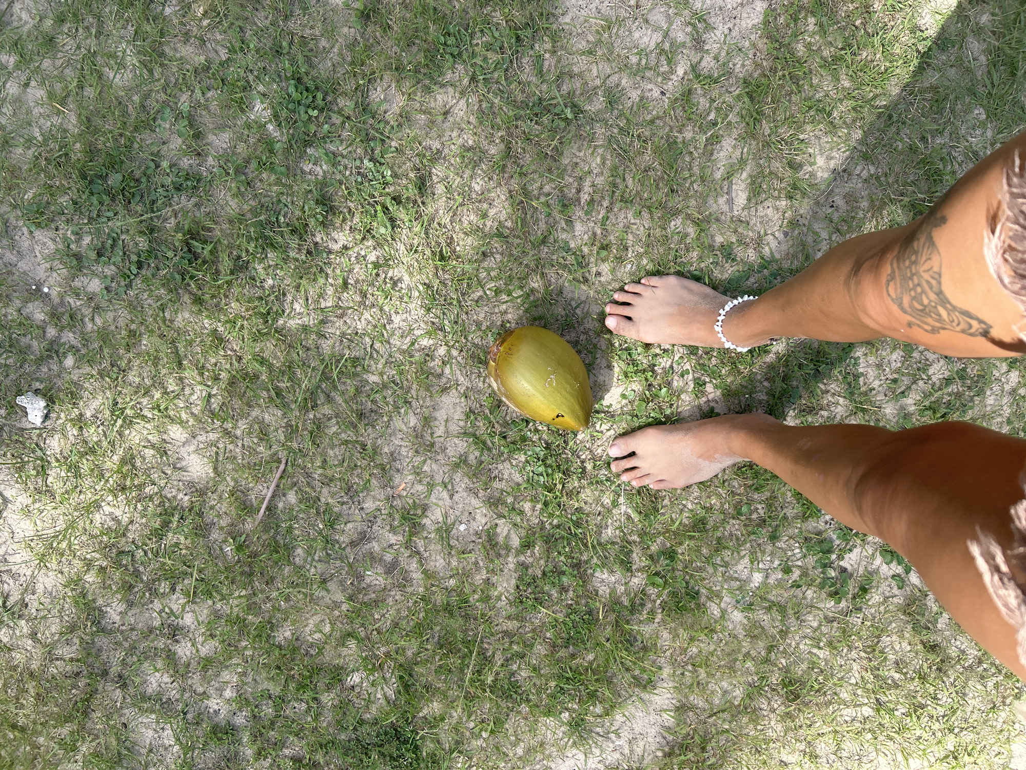 The image shows a person's feet standing on a patch of sandy ground with sparse green grass and a few small plants. A single green coconut is positioned between the person's feet. The person has a tattoo on their right thigh and is wearing a white beaded anklet on their left ankle. The ground appears to be a natural outdoor setting, possibly a beach or park area in Ban Khlong Hia, Thailand, given the presence of sand and vegetation. The lighting suggests it is daytime.