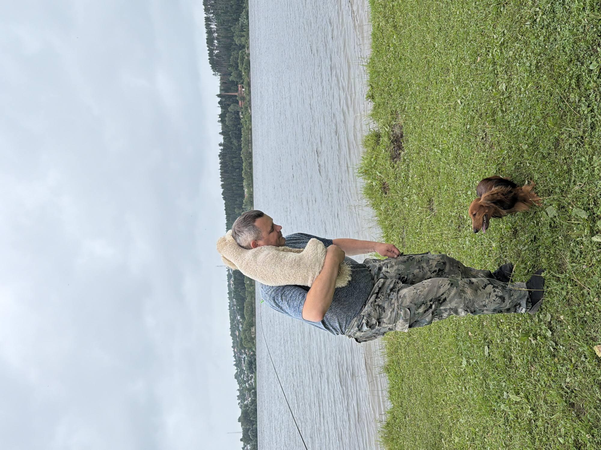 A man is carrying a fluffy stuffed animal over his shoulder near a body of water. He is wearing a blue t-shirt and camouflage pants. A small brown dog is standing on the grass in front of him, looking up. The man is looking down at the dog. In the background, there is a body of water with a fishing line in it, a tree line, and a town with buildings. The sky is cloudy. This scene is likely along the Kama River in Chaikovsky, Russia.