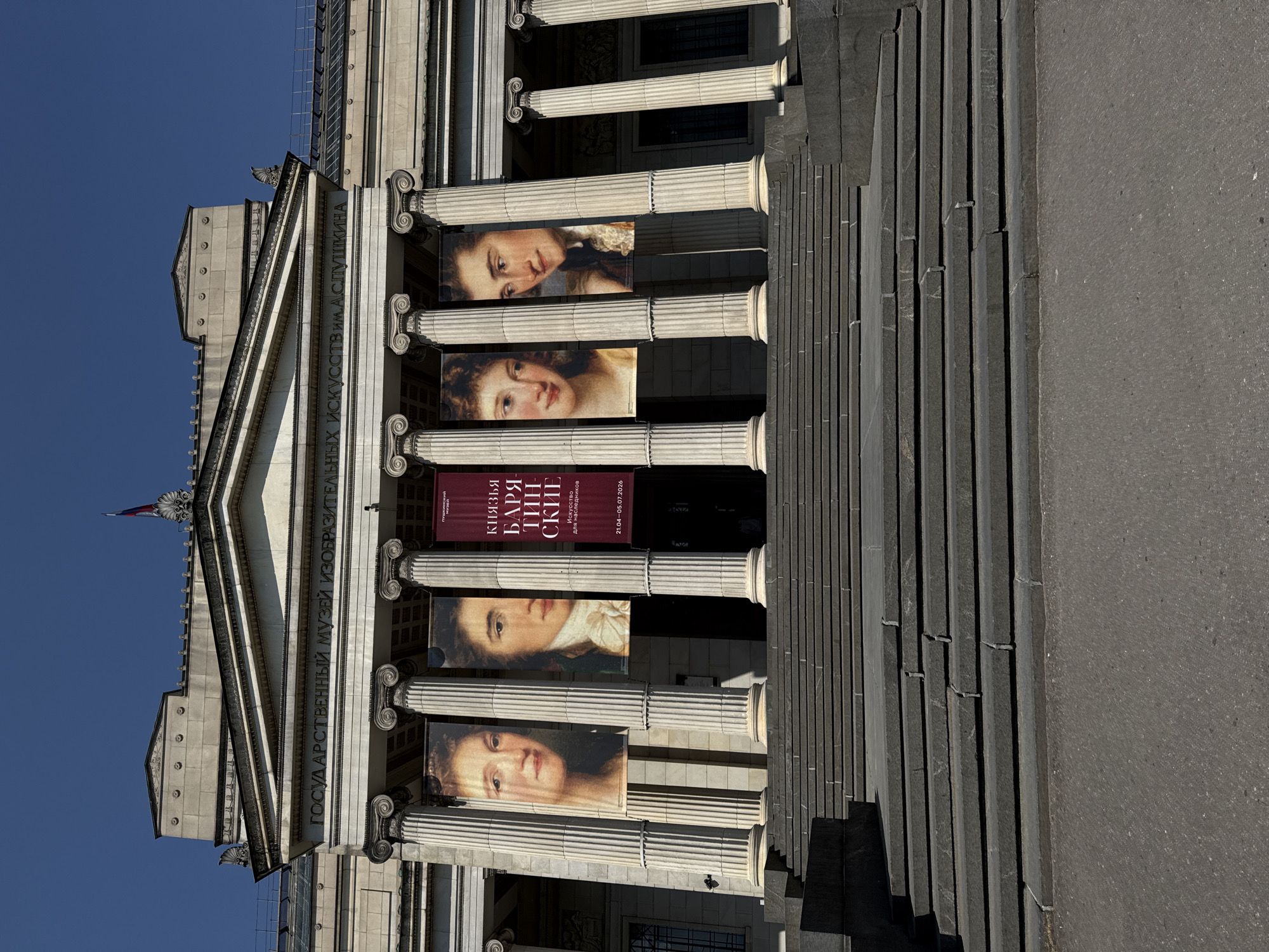 The image depicts the facade of the Pushkin State Museum of Fine Arts in Moscow, Russia. The building has a classical architectural style with prominent columns. Banners featuring portraits and promotional text related to the "Princes Baryatinsky" exhibition are displayed between the columns. The foreground includes a set of stone stairs leading up to the building. The sky is clear and blue.
