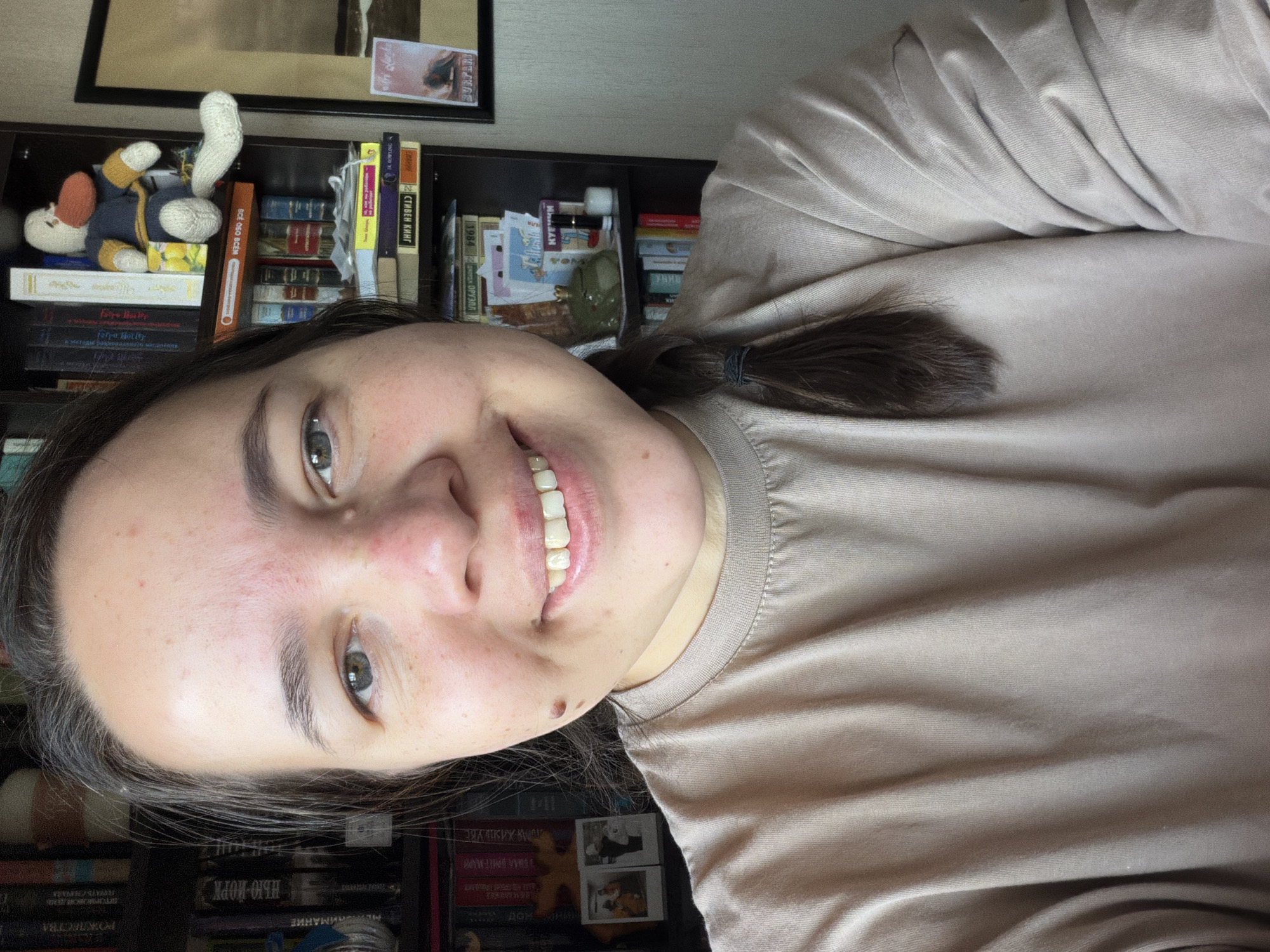 A woman is smiling at the camera. Behind her, there is a bookshelf filled with books and other objects. The books have Russian titles on them: "РОЖДЕССТВА", "НЬЮ-ЙОРК", "GETOPMOBOЙДЕНЬ", "ПОЧАТЬ СНАЧА", "СТОРИЯ ЖИЗНЬ УВЕ", "fatya Hotity", "W308 090 309", "ДЖОРДЖ ОРУЭЛЛ", "СТИВЕН КИНГ", "ROWLING". Other objects on the shelf include a stuffed animal and various decorative items. The image was taken in the city of Chaikovsky, Russia.
