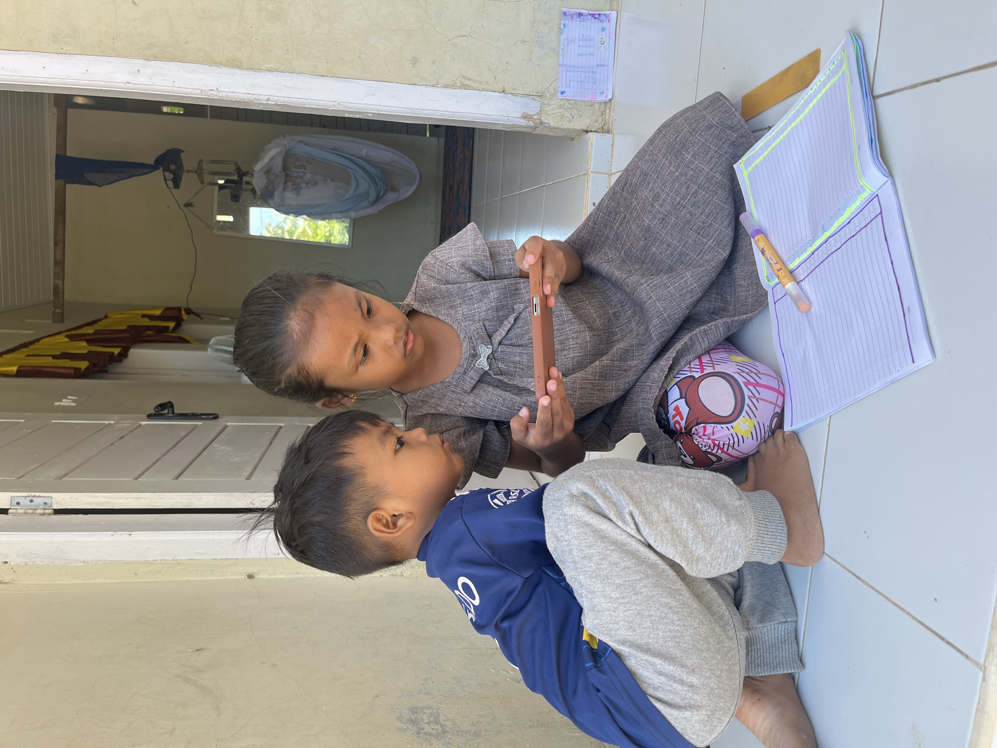 Two young children, a boy and a girl, are sitting on a tiled floor outdoors in front of a doorway. The boy, on the left, is wearing a blue t-shirt with white lettering and grey sweatpants. He is looking intently at a smartphone that the girl is holding. The girl, on the right, is wearing a grey dress and is focused on the smartphone as well. In front of them are an open notebook with colorful borders, a pen, a ruler, and a colorful, cartoon-themed ball. The background shows an open doorway leading into a dimly lit interior with a hammock visible. The exterior wall is textured and painted a light color. The scene appears to be in a residential area in Geunteng, Indonesia, based on the provided context.