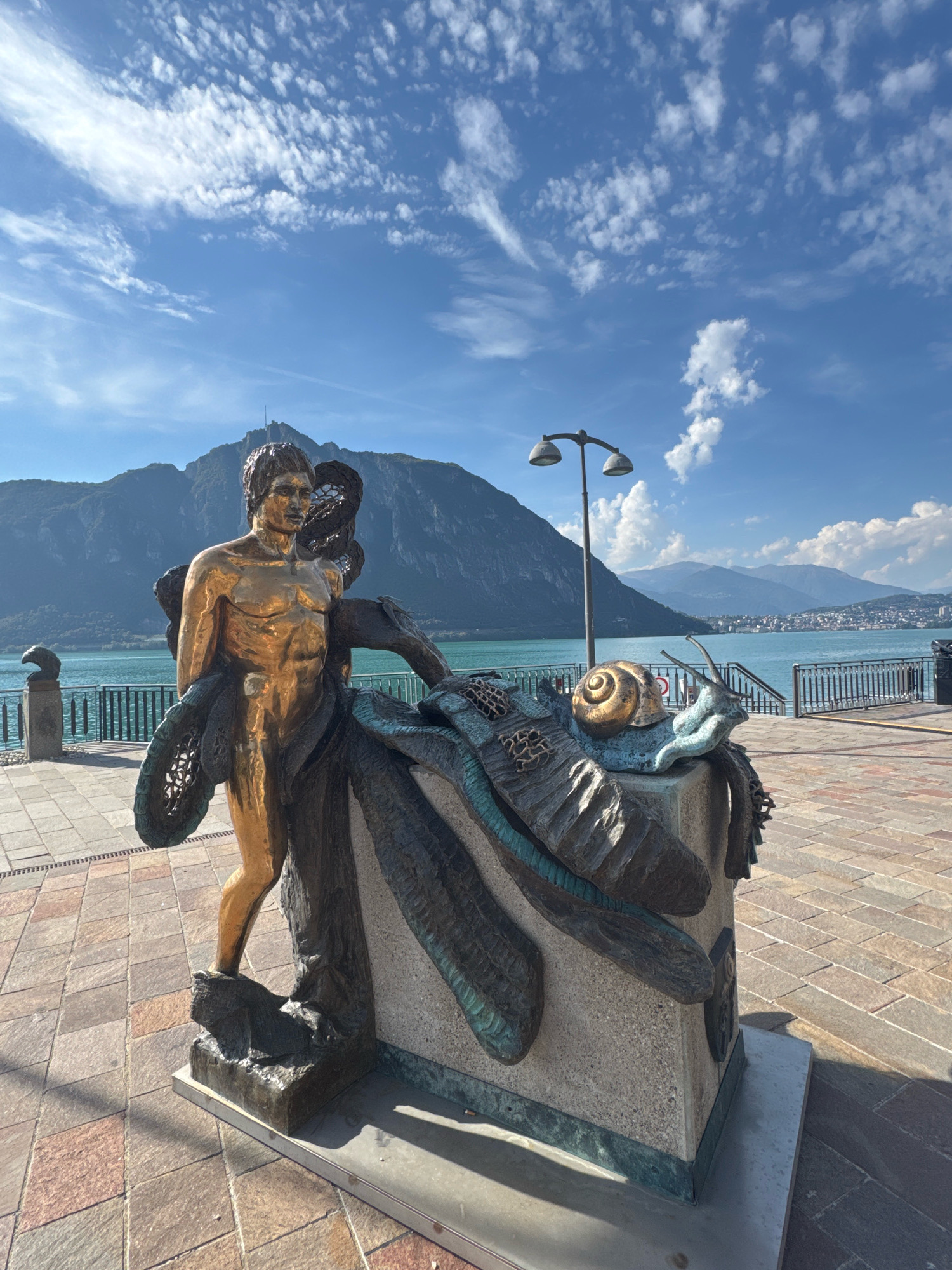 A golden statue of a muscular man stands to the left of a bronze sculpture of a snail. The man is partially draped in what appears to be a dark, textured fabric or material that also wraps around the snail sculpture. The snail sculpture is on a light-colored, rough-textured pedestal. The scene is outdoors, paved with stone tiles in a diamond pattern. In the background, a wide body of water, likely a lake, stretches across the frame. Beyond the water, a cityscape with buildings is visible, nestled at the base of large, imposing mountains. The sky above is a vibrant blue, dotted with wispy white clouds. A lamppost with two lights stands behind and to the right of the man's statue, near the edge of the water. A dark metal railing runs along the water's edge. The image is taken from a low angle, emphasizing the statues and the expansive mountain and sky scenery. This is a view of the lakefront promenade in Campione d'Italia, Italy.