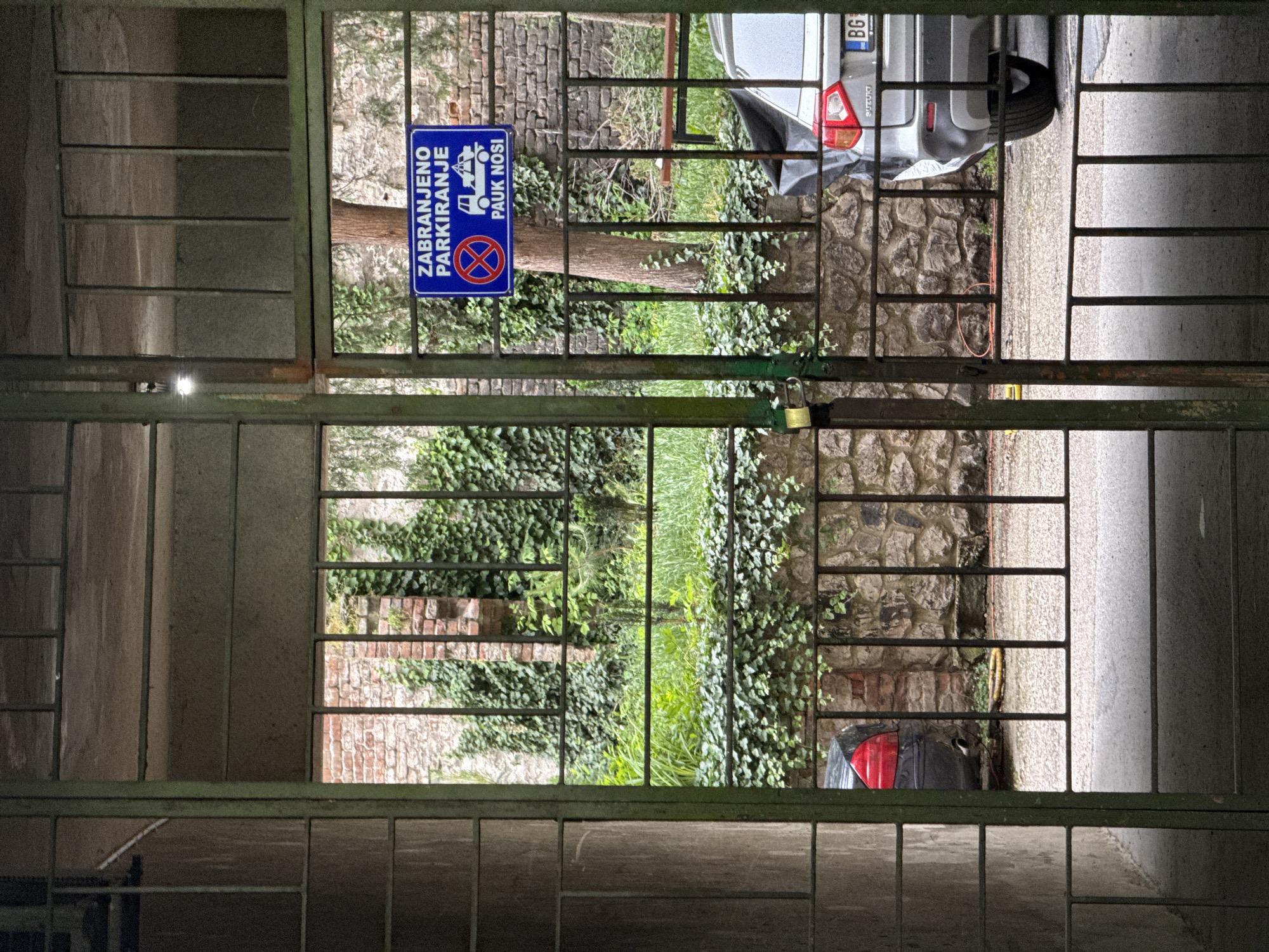 Through a metal grid gate, a parking area is visible in Belgrade, Serbia. A no parking sign ("Zabranjeno parkiranje; pauk nosi") is posted on the left. A silver Suzuki car, with license plate "BG", is parked in the area. The area is paved and bordered by brick and stone walls overgrown with ivy.