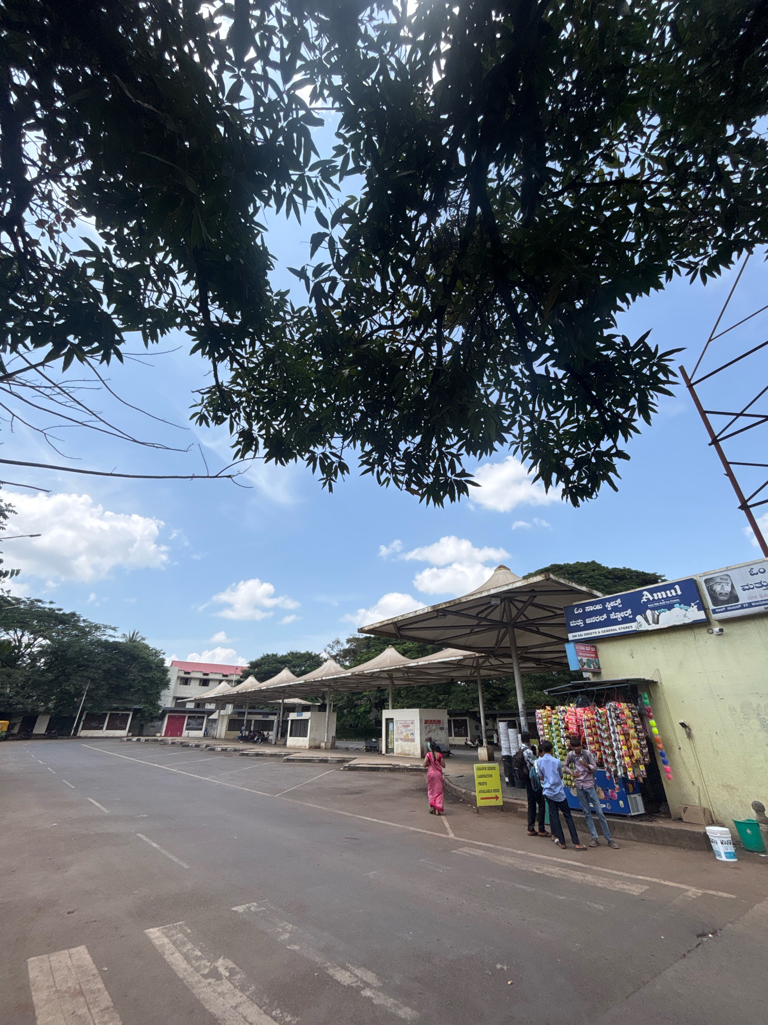 The image captures a street scene with a bus stop or waiting area on the right side, under a canopy supported by white pillars. Several people are standing near a small shop attached to the bus stop, which is adorned with colorful packets of snacks and other goods. A sign for "Amul" is visible on the shop's facade, suggesting it sells dairy products and snacks. In the foreground, an asphalt road with white lane markings is visible. Trees with dense foliage overhang the scene from the top. In the background, a multi-story building with a red roof can be seen, along with more trees and a bright blue sky with scattered clouds. The overall impression is a typical daytime scene in an Indian city, likely in a somewhat commercial or transit area.
