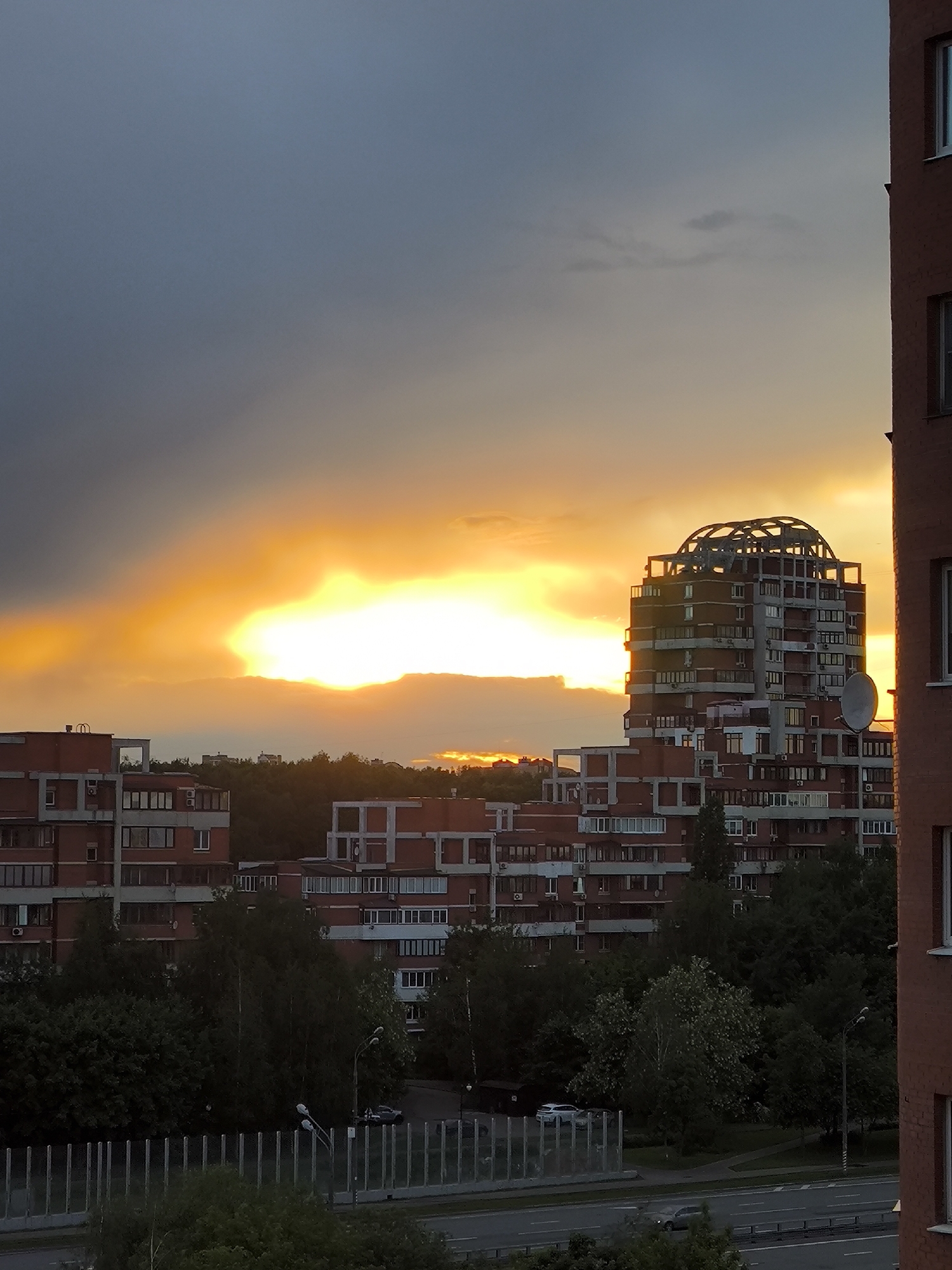 The image shows a sunset over a city, likely located in Russia given the architectural style of the buildings. The sky is predominantly grey and orange, with a bright yellow patch where the sun is setting behind a layer of clouds. Several red brick buildings of varying heights dominate the skyline. The tallest building is of particular interest due to its unfinished dome-like structure on top. Trees are seen filling the spaces between the buildings, adding a touch of greenery to the urban scene. In the foreground, there is a road with a few cars on it, along with a fence-like structure possibly to protect the street from noise.