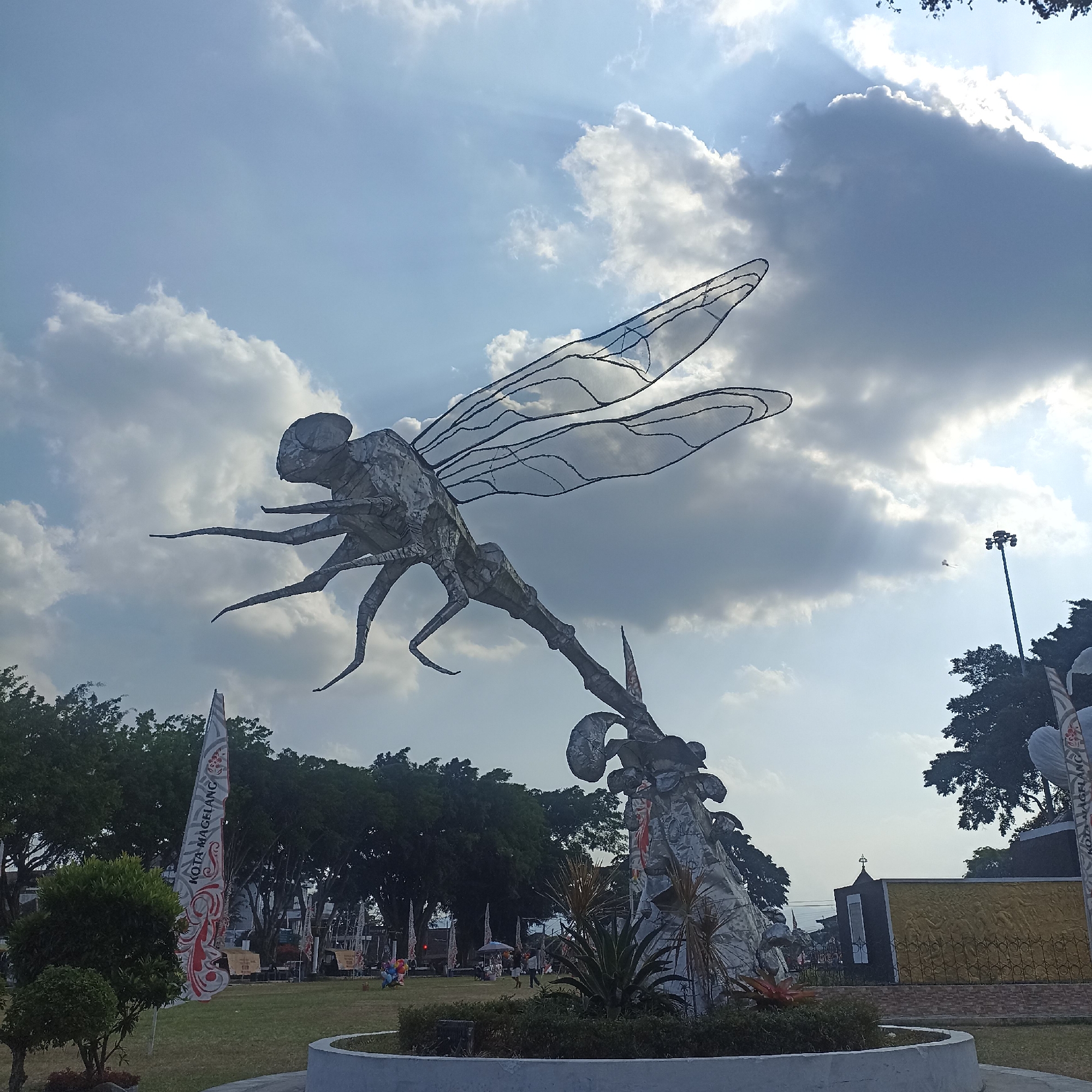 A large, metallic sculpture of a dragonfly is the central focus of this image, positioned outdoors in a park-like setting. The dragonfly sculpture is in flight, with its intricate, wireframe wings spread wide and its segmented body and legs clearly visible. It appears to be constructed from a weathered, silvery material, possibly metal sheets or panels. The sculpture is mounted on a textured, rocky base, which is surrounded by a low, circular concrete planter containing some greenery.
In the background, a bright, partly cloudy sky dominates the upper portion of the frame, with sunlight filtering through the clouds in visible rays. Below the sky, a line of trees forms a green backdrop. To the left, a tall, striped flag with red and white patterns and text that reads "KOTA MAGELANG" (City of Magelang) is visible. There are other flags and structures in the distance, suggesting a public event or recreational area.
A few scattered people are visible in the mid-ground and background, engaged in various activities like walking and sitting. Some small trees and bushes are present in the foreground and mid-ground, as well as a lamppost on the right side of the image. The overall environment appears to be a well-maintained urban park in the city of Magelang, Indonesia, likely a public space with decorative elements and recreational amenities. The time of day appears to be late afternoon or early evening, given the angle of the sunlight and the quality of the light.