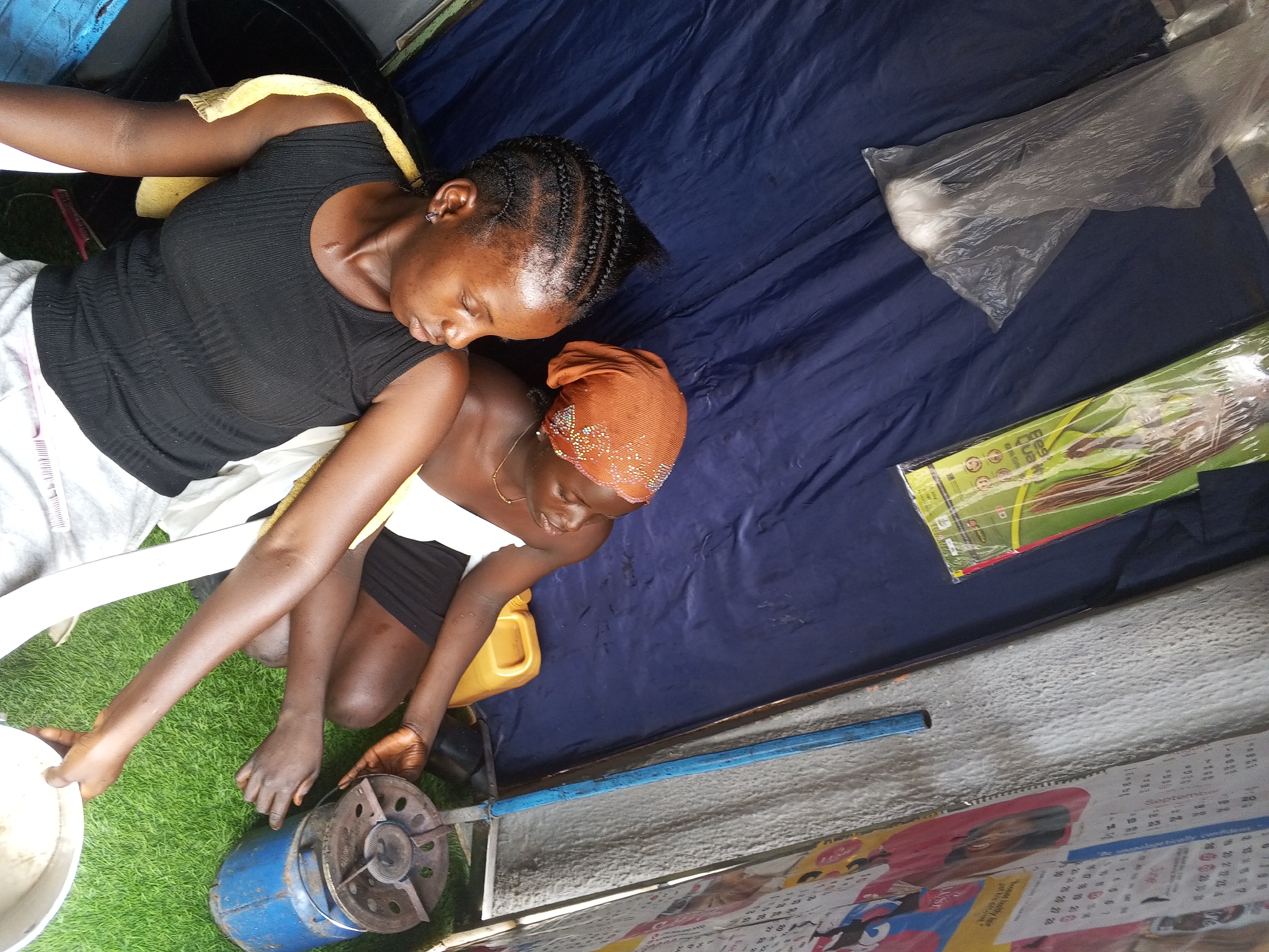Two women are positioned outdoors beside an old blue propane gas stove. One woman, with braided hair, is seated on a white plastic chair wearing a black top and white shorts. She is holding a white pot or bowl. The other woman is kneeling and wearing an orange headscarf, a white tank top, and a black skirt. A yellow plastic container is nearby. A blue tarp is laid out, and a plastic bag, a package of items, and a blue metal pipe are visible on the tarp. A calendar is attached to a wall nearby. The setting appears to be a modest outdoor cooking area.