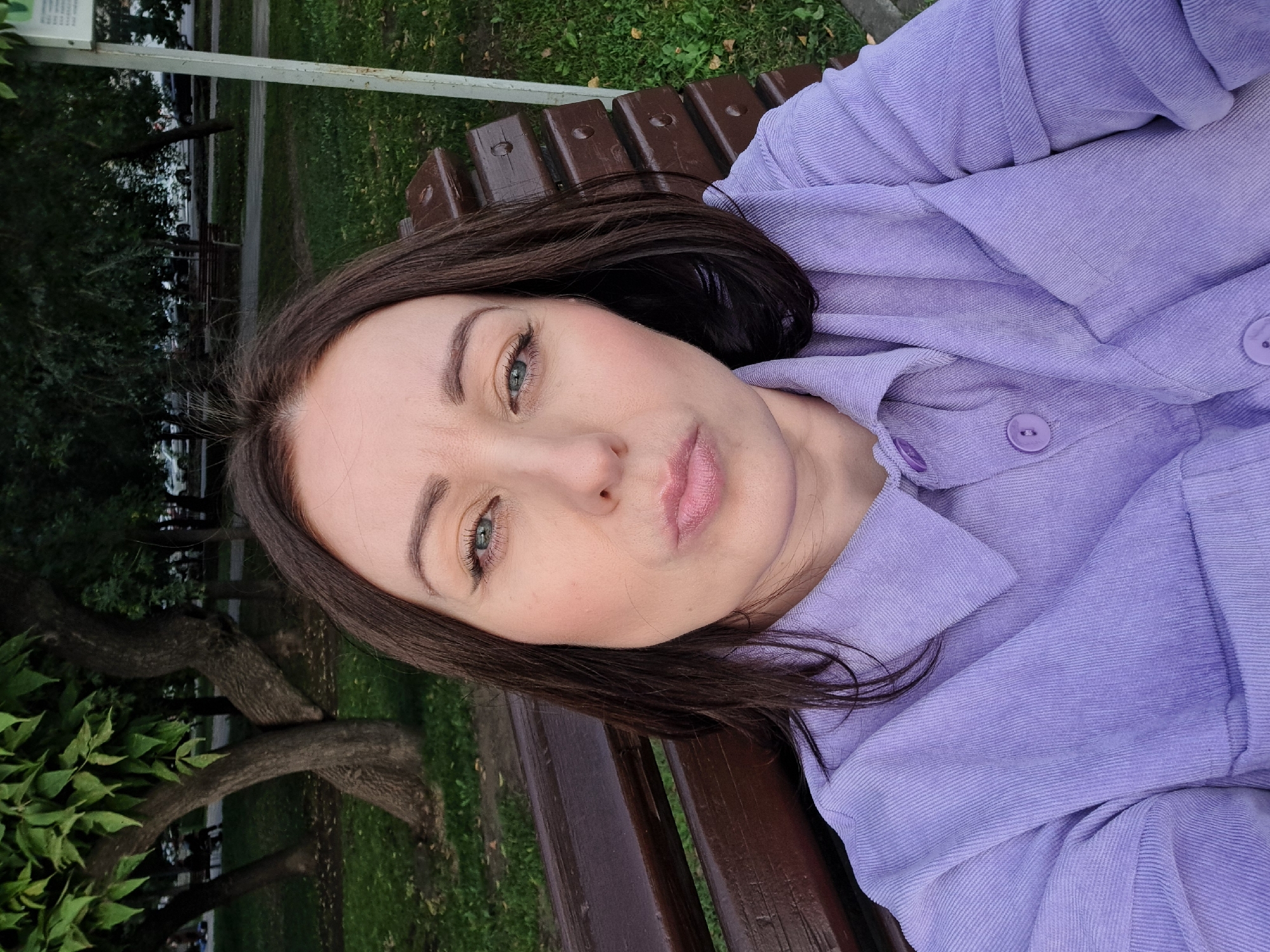 A woman with dark brown hair, wearing a purple corduroy shirt, is sitting on a wooden park bench. She is looking at the camera with a playful expression, puckering her lips. The background shows trees, grass, and a street with cars. The image appears to be a selfie taken outdoors. The presence of trees and grass suggests a park setting, and the cars on the street indicate an urban environment. The season seems to be either late spring or summer, given the foliage on the trees. The woman is wearing makeup, including eyeliner and lipstick. She is sitting on a bench with a slatted wooden seat and backrest. The overall scene is casual and informal. There is no specific indication of the location being Irkutsk, Russia, or any other specific city.