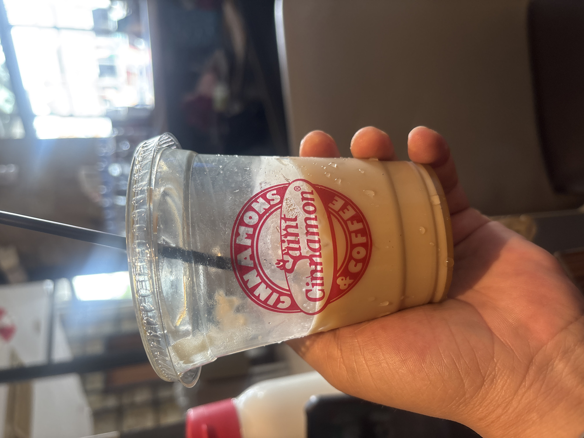 A hand holds a clear plastic cup filled with a light brown liquid, likely a coffee drink, with condensation on the exterior. A black straw is inserted into the cup. The cup has a red and white logo that reads "CINNAMONSaint Cinnamon & COFFEE." In the background, out of focus, a white bottle with a red cap and other blurry objects are visible. The environment appears to be indoors, possibly a cafe or restaurant, with natural light streaming in from windows. The image is framed from a close-up perspective, focusing on the cup and the hand holding it. No specific location in Pekanbaru, Indonesia is discernible from the image itself.