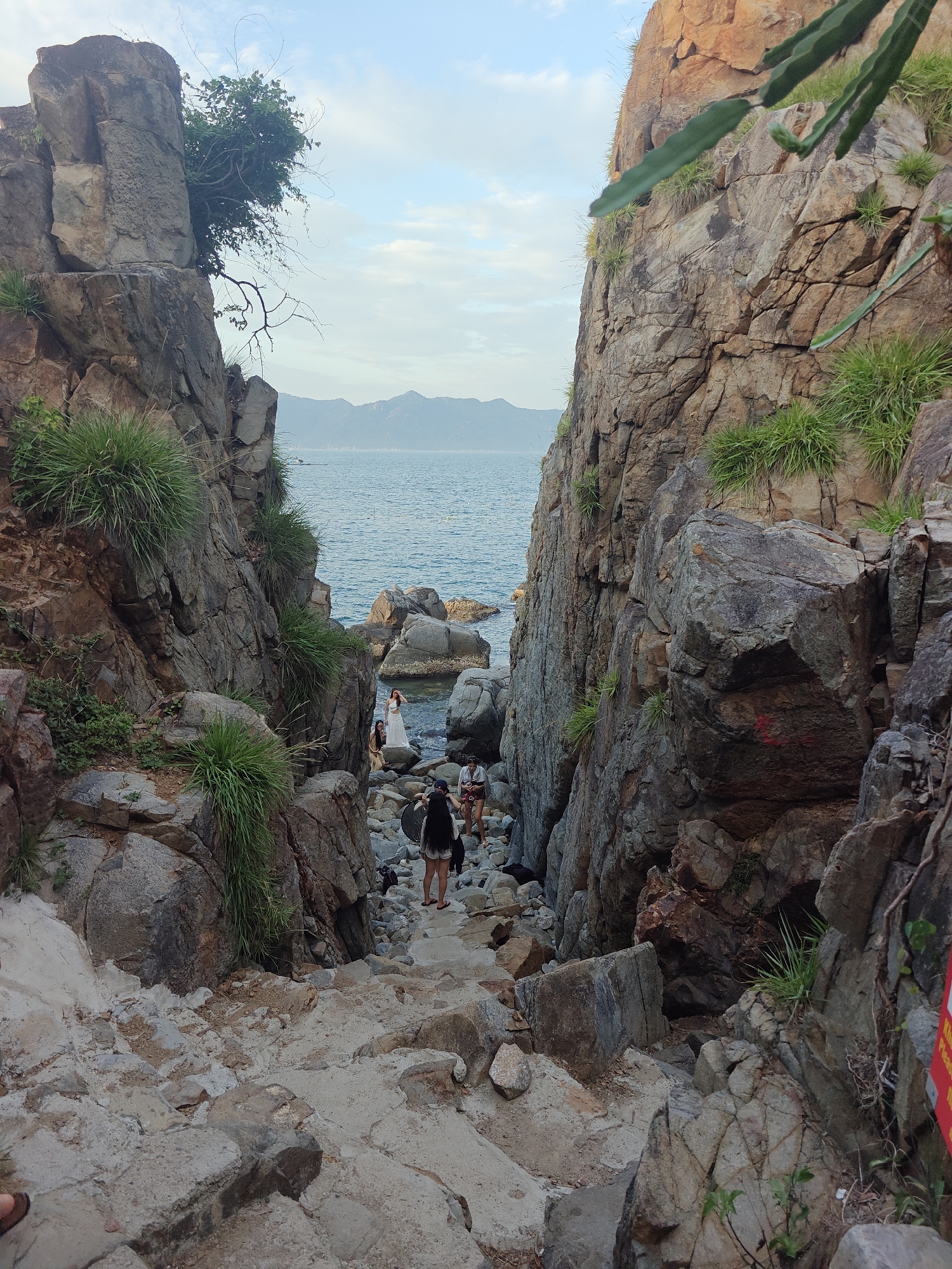 The image depicts a rocky, coastal pathway leading down towards the sea in Vĩnh Lương, Vietnam. The pathway is flanked by large, rugged rock formations covered with sparse green vegetation. In the distance, the blue sea stretches out to the horizon, meeting a hazy sky with distant mountains visible. A group of people are visible on the path and near the water's edge. Two women, one in a white dress, appear to be posing for photos on the rocks by the sea. Further down the path, several other individuals are walking or standing. The overall scene conveys a natural, somewhat untamed coastal environment with human activity.