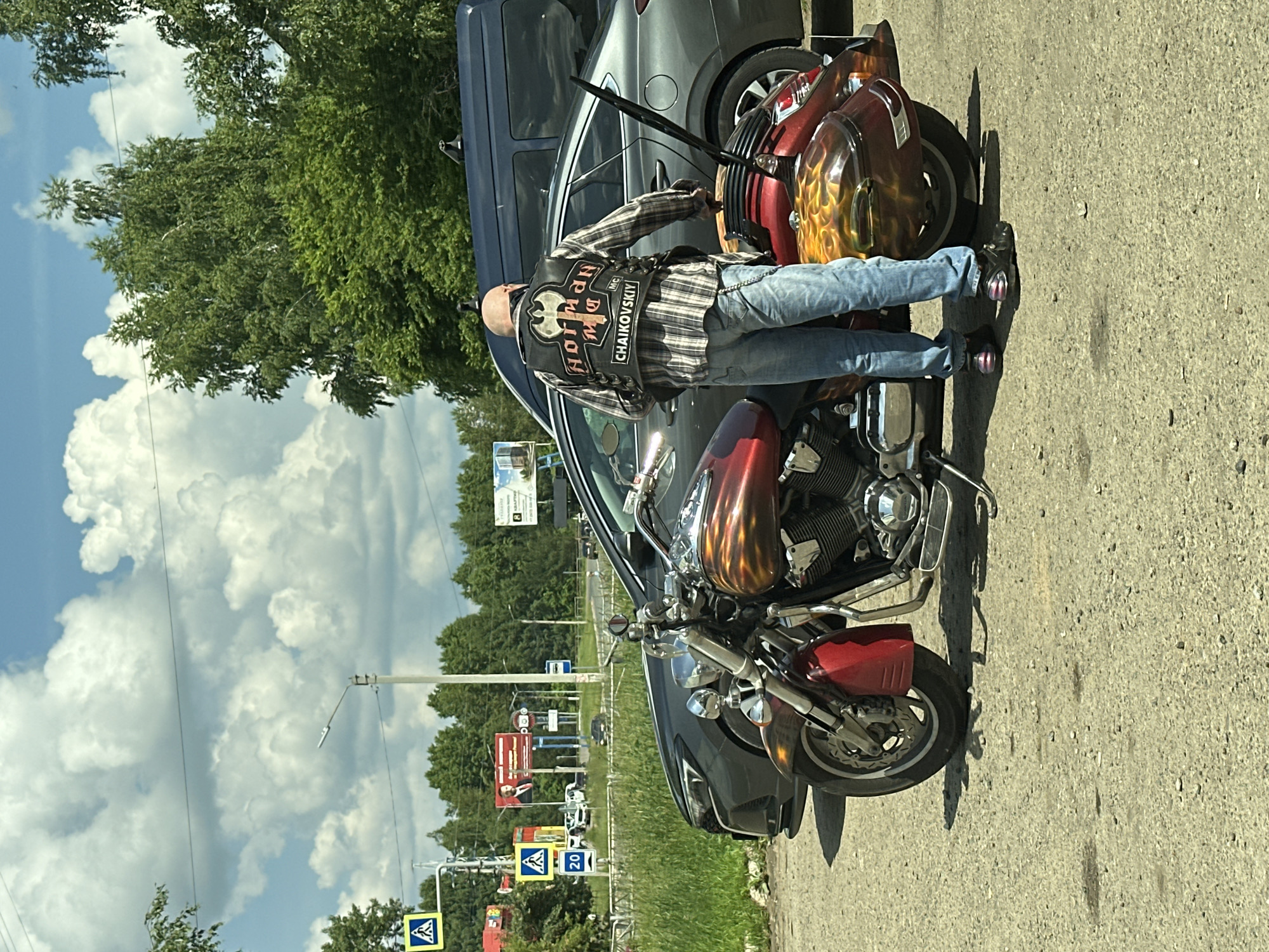 A bald man, wearing a checkered shirt and a leather vest with patches, stands next to a red and chrome motorcycle with flame graphics. The vest has the text "NORMAN MC CHAIKOVSKIY" and an axe emblem. The man is wearing blue jeans and black shoes with reflective accents. The motorcycle is parked on a gravel surface next to a dark-colored sedan and a dark-colored van. In the background, there are trees, traffic signs, and billboards, suggesting an urban or suburban environment. One billboard features a portrait of a man in a suit. Another billboard displays text in Cyrillic, with one section appearing to be an advertisement for real estate. The sky is partly cloudy. The overall scene appears to be a casual stop, possibly in a parking area or along a roadside in the city of Chaikovsky, Russia.