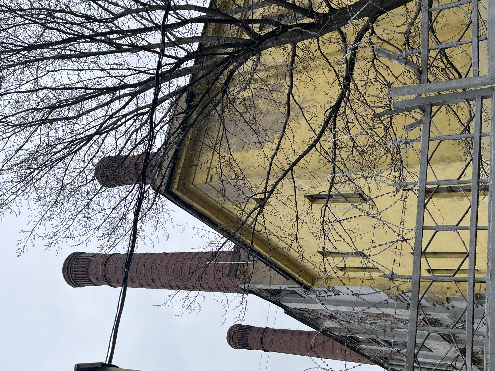 An urban or industrial landscape is depicted from a low, upward-looking perspective, with the image rotated 90 degrees counter-clockwise. The scene is dominated by three tall, reddish-brown brick industrial chimneys and a large, faded yellow building, all partially obscured by a dense network of bare, leafless tree branches.
On the left side, two cylindrical brick chimneys rise against an overcast gray sky. The chimney higher on the left is taller, showcasing detailed brickwork at its cap. A second, slightly shorter chimney is situated below it. Dark electrical wires or cables run diagonally across this sky and tree-filled area.
To the right, a dilapidated building with a faded and peeling yellow facade occupies much of the frame. It features architectural elements like ledges or cornices, and hints of windows are visible. The bare tree branches extend across the sky and in front of the yellow building, creating an intricate, web-like pattern.
In the lower right portion of the image, a third, shorter brick chimney is partially visible. Further to the right, a gray metal structure, possibly a ladder or part of a fence, stands vertically. This structure is entangled with strands of barbed wire that also stretch across the bare branches and in front of the yellow building.
The scene conveys a sense of age and disuse, typical of an old industrial area, possibly in winter or early spring due to the bare trees. The overcast sky and the elements of decay contribute to a stark and somewhat somber mood. There are no people visible, and no discernible activity or text.