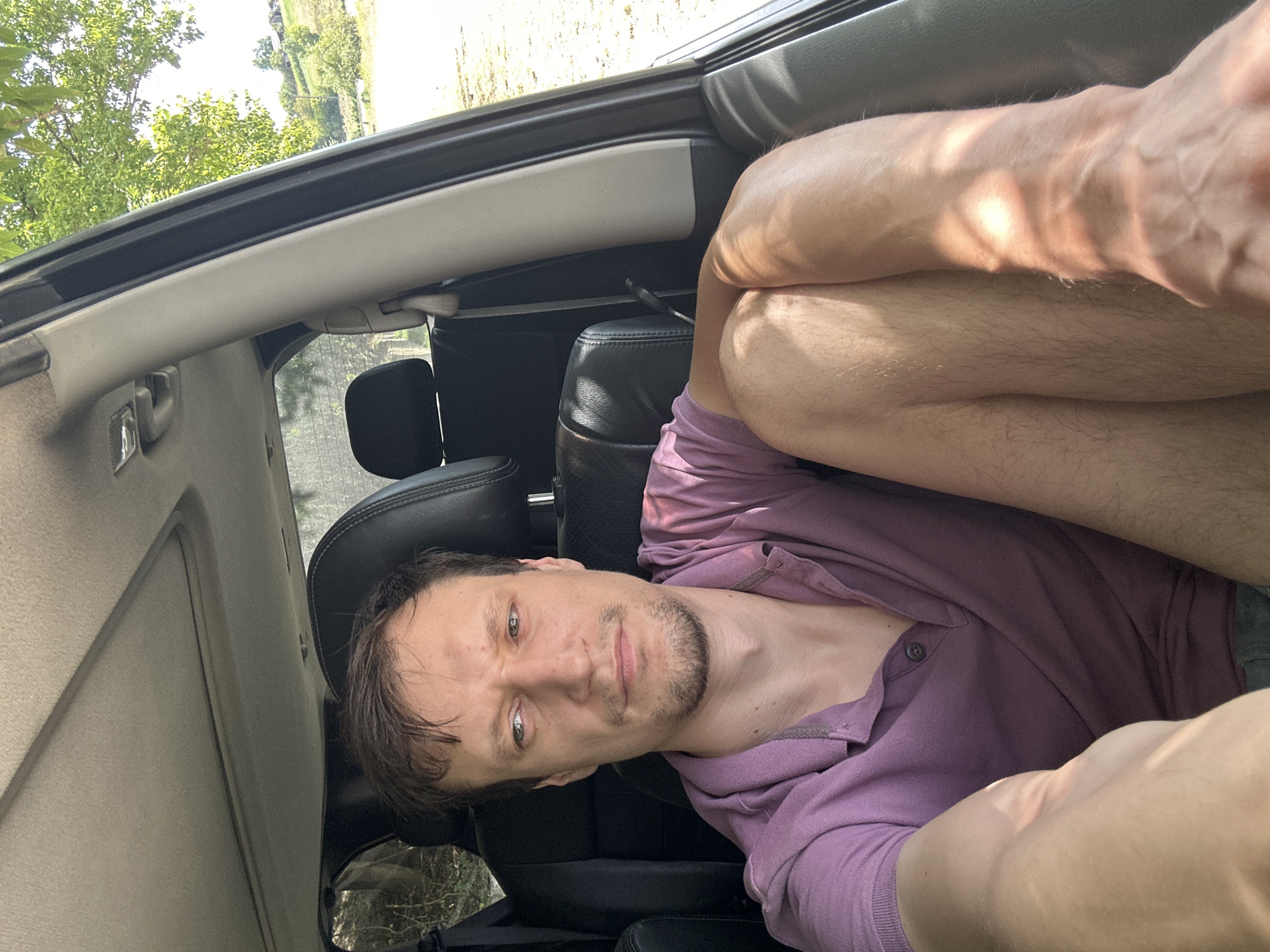 A medium shot captures a man from the chest up, seated inside a vehicle. He is wearing a purple polo shirt and has short brown hair and stubble. His expression is neutral, and his eyes are looking slightly to his left. Sunlight streams through the car's windows, casting shadows on his skin and clothes. The interior of the car is visible, including the seats and the sunroof. The background outside the car shows a sunny, rural landscape with trees and rolling hills, consistent with the countryside around Makiv, Ukraine. The overall scene appears to be a candid, outdoor photograph taken during the daytime.