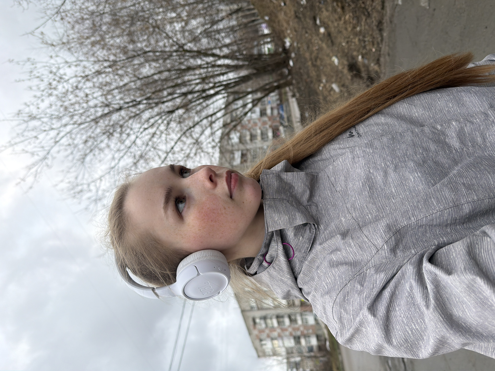 A fair-skinned adolescent female with freckles is the primary subject. She has long, straight, light brown hair partially pulled back, and is wearing white headphones. She is dressed in a heather grey jacket. The girl is glancing upwards and to the left.
The background features a street scene in the city of Berezniki, Russia. Bare deciduous trees line the street, with a grey apartment building visible in the distance. Patches of dried grass and debris are scattered along the roadside. The sky is overcast.