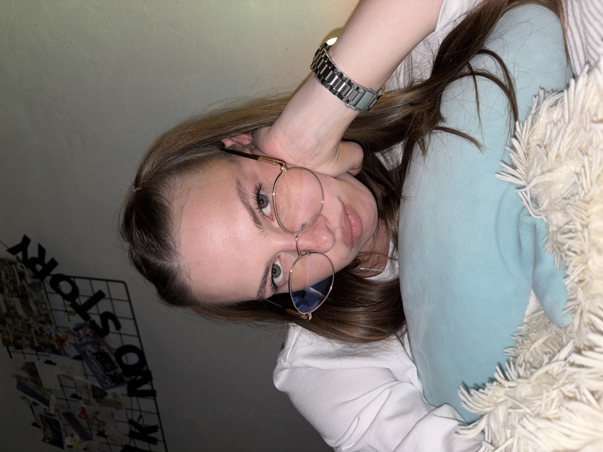 A young woman with long brown hair, wearing glasses and a white shirt, rests her head on a light blue pillow. She is lying down, with her right arm supporting her head. A metallic watch is visible on her wrist. In the background, a wire grid with pictures and black letters spelling "K NO STORY" is attached to a wall. The lighting suggests an indoor environment, possibly a bedroom, with warm light casting shadows on the wall. The scene appears to be a selfie taken in Perm, Russia, based on the implied context of the prompt.