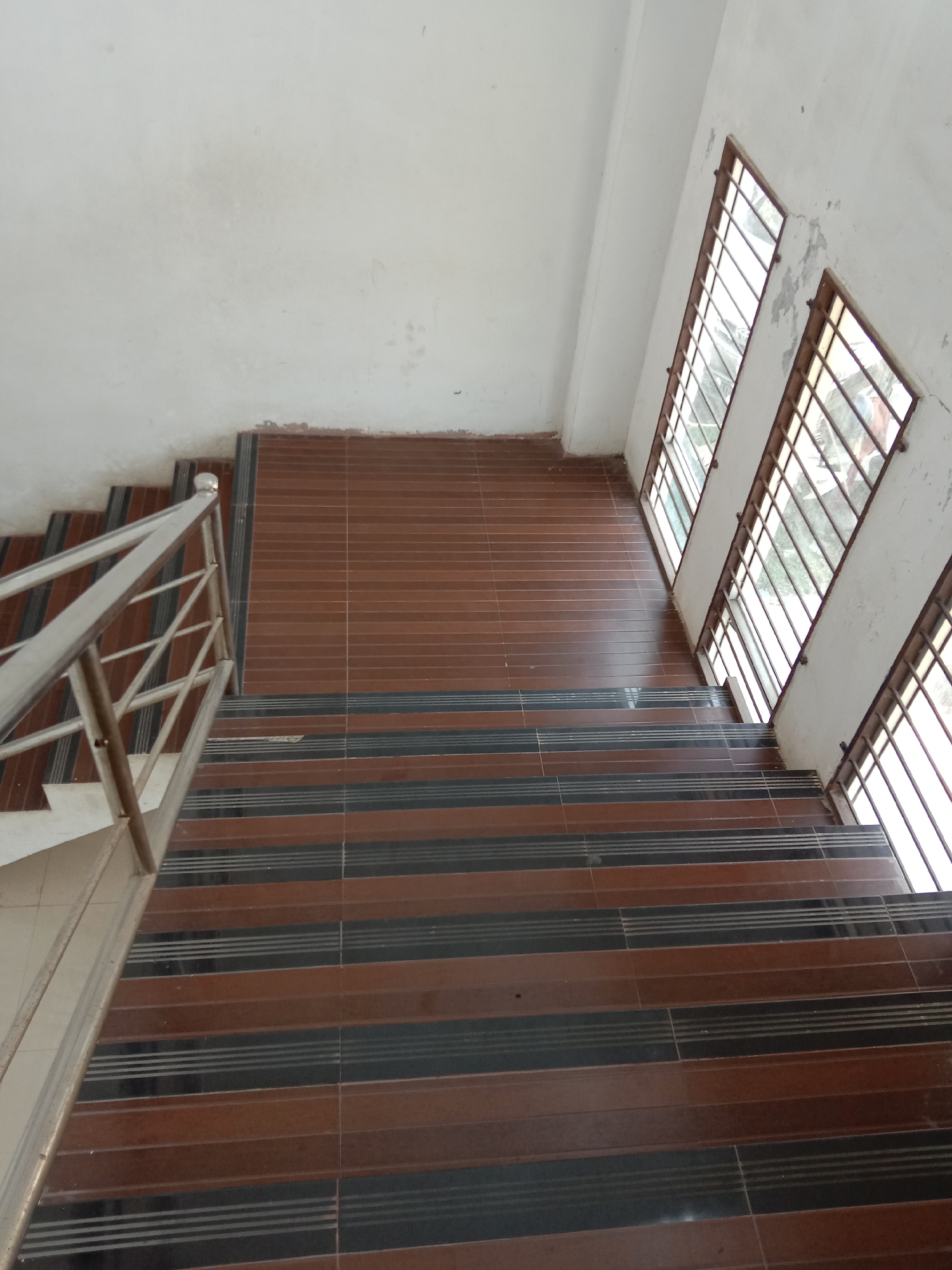 The image displays an interior view of a staircase and an adjacent landing in a building, captured from a high-angle perspective looking downwards. The staircase features steps with a distinct alternating pattern of dark brown and black rectangular tiles. The black tiles incorporate several thin, light-colored horizontal lines, possibly for grip or decoration. A metal handrail runs along the left side of the staircase, made of polished silver-colored material, likely stainless steel.
To the right of the staircase, a white or off-white wall houses three tall, narrow windows. These windows are fitted with metal security bars, forming a grid pattern. Natural light streams through the windows, illuminating the scene, indicating it is daytime. The walls show signs of wear and age, with visible discoloration, dirt, and some peeling paint, particularly around the window frames. The landing area at the top of the visible stairs is also tiled with similar brown rectangular tiles. No people or specific activities are visible in the static scene. There is no visible text. The overall appearance suggests a functional, possibly public, space in Lhokseumawe, Indonesia.