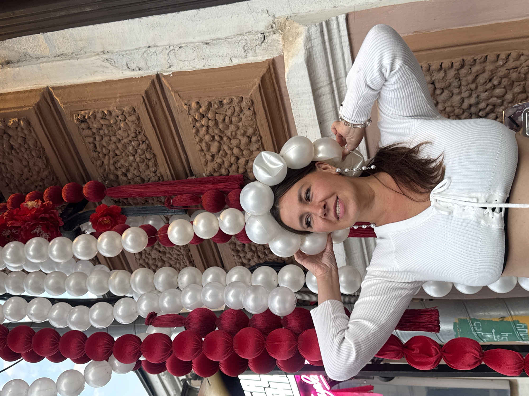 A woman with brown hair and fair skin, wearing a white ribbed top and pearl earrings, stands outdoors in front of a building with ornate architectural details. She is holding a headpiece made of large white spherical ornaments, resembling a crown or halo, and her left hand is touching the ornaments. Behind her, strings of red and white spherical ornaments hang vertically, some appearing to be made of velvet and others of a glossy material. Red tassels are also visible among the decorations. The background includes a textured building facade and what appears to be part of a street sign or advertisement. The scene is likely outdoors in an urban setting, possibly during a festive event.
