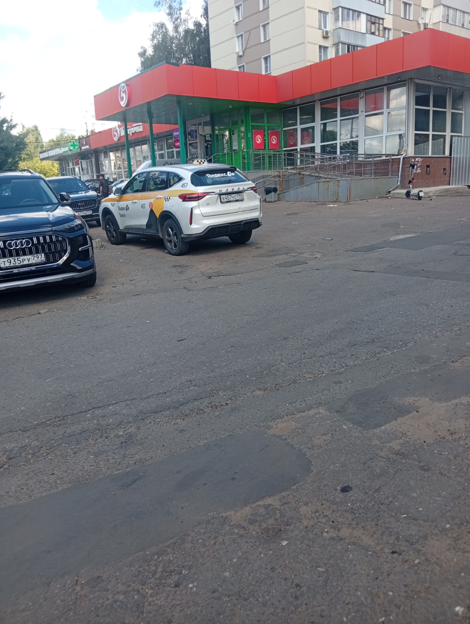 The image shows a street scene in Pushkino, Russia, in front of a grocery store with a red and green facade. A white Yandex.Go taxi is parked on the asphalt, with a dark-colored Audi in the foreground. The taxi has a Russian license plate that reads "X457YC 750". The Audi's license plate reads "T935PY 797". There are no people clearly visible in the scene. The environment includes a sidewalk, a ramp leading to the store entrance, and trees in the background. The ground is paved with asphalt and shows signs of wear and tear.