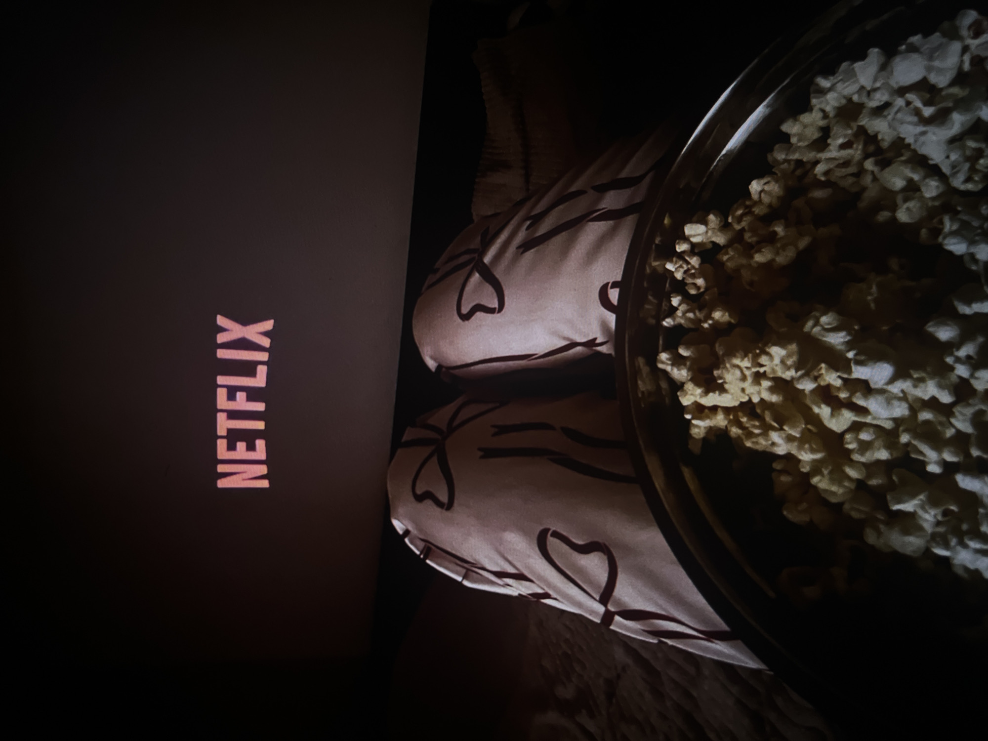 The image shows a screen displaying the Netflix logo, with a person wearing pajamas with a heart pattern on their legs. A large bowl of popcorn is visible next to the person. The lighting is dim, suggesting a nighttime environment indoors. The location is implied but not specified, with the city being described as Unknown.
