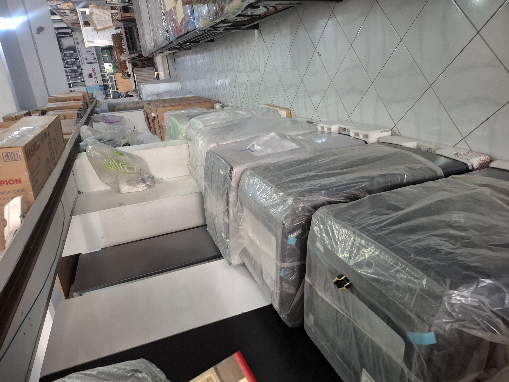 The image depicts the interior of a retail store, likely a furniture or appliance shop, located in Banjarmasin, Indonesia. The store features rows of merchandise, predominantly appliances wrapped in clear plastic sheeting, such as washing machines or refrigerators, arranged along a tiled floor. In the background, there are shelves stocked with boxes, and some furniture pieces, including a bed frame with mattresses and a decorative headboard. A ceiling fan is visible hanging from the ceiling. The overall impression is one of a commercial establishment displaying a variety of goods for sale.