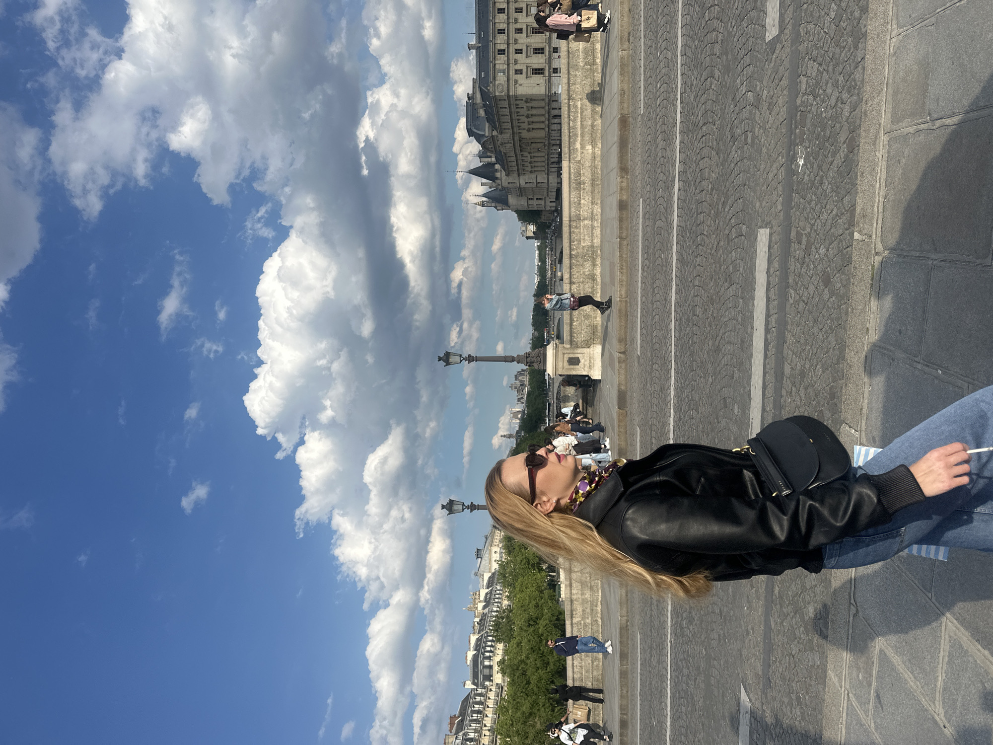 A woman with long blonde hair is standing on a cobblestone street in Paris, France. She is wearing sunglasses, a black leather jacket, blue jeans, and a black purse. She is holding a cigarette in her right hand. The sky is blue with puffy white clouds. Buildings and lamp posts can be seen in the background. People are walking along the bridge. The building in the background is the Conciergerie.