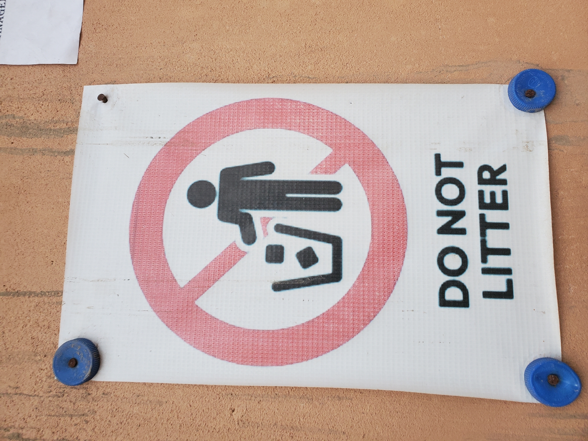 A "DO NOT LITTER" sign is affixed to a textured, light brown wall in Bukuru, Nigeria. The sign is white with a red circle and slash symbol over a black silhouette of a person dropping an item into a trash bin. Below the symbol, the words "DO NOT LITTER" are printed in bold black capital letters. The sign is held in place by four blue plastic bottle caps, each fastened with a nail. The wall appears to be exterior, possibly of a building, with some faint vertical streaks of dirt.