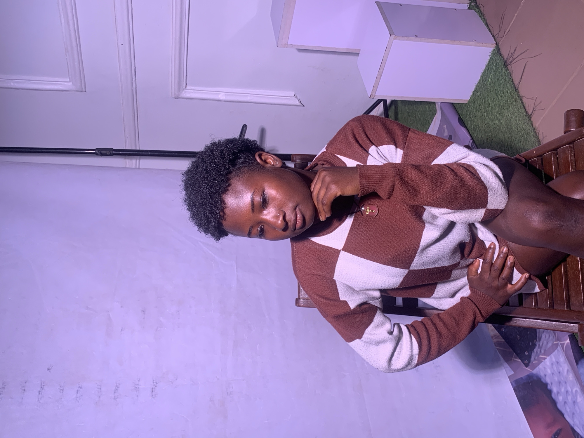 The image shows a young Black person sitting in a wooden chair. They are wearing a brown and white checkered sweater and have short, curly black hair. Their head is tilted down, and they are looking to the side with a thoughtful expression. Their left hand is resting on their chest, near a small gold pendant necklace, and their right hand is resting on their knee.
The background is a light purple wall with some decorative molding at the top. To the right of the person, there are some white geometric shelves and a patch of artificial grass. The lighting in the image is dim and has a purple tint, suggesting it might be evening or taken indoors with artificial lighting. The overall scene appears to be a casual portrait session or a candid moment captured in a studio-like setting.
There is no visible text in the image. The location context is Barakin Akawo, Nigeria.