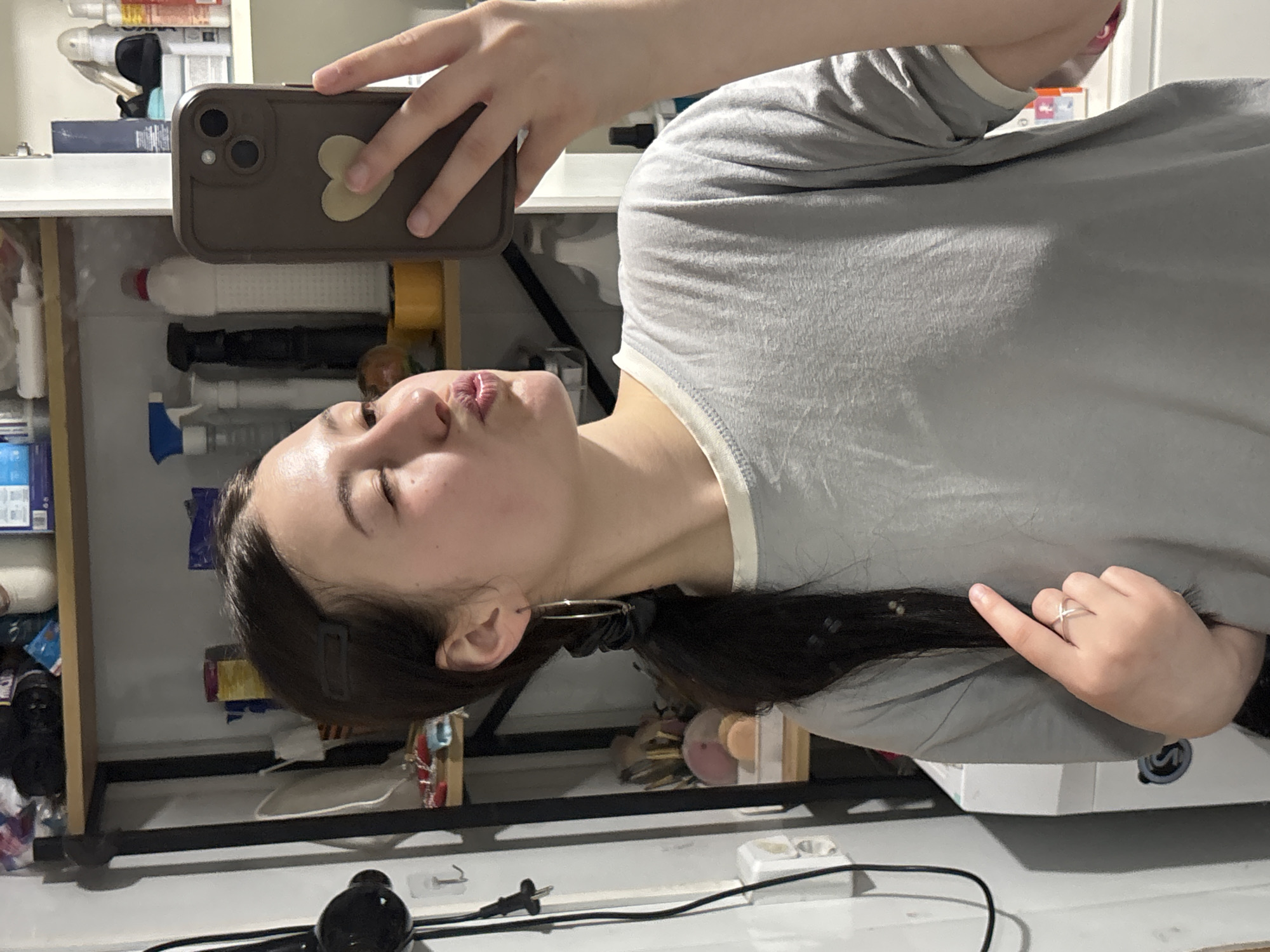 A woman with dark hair and fair skin is taking a selfie with a brown cell phone. She is wearing a gray t-shirt and has an earring in her left ear. Her lips are pursed as she takes the photo. The background shows a cluttered shelf with various bottles and containers. A black power cord and plug are on the white floor. The location cannot be determined.