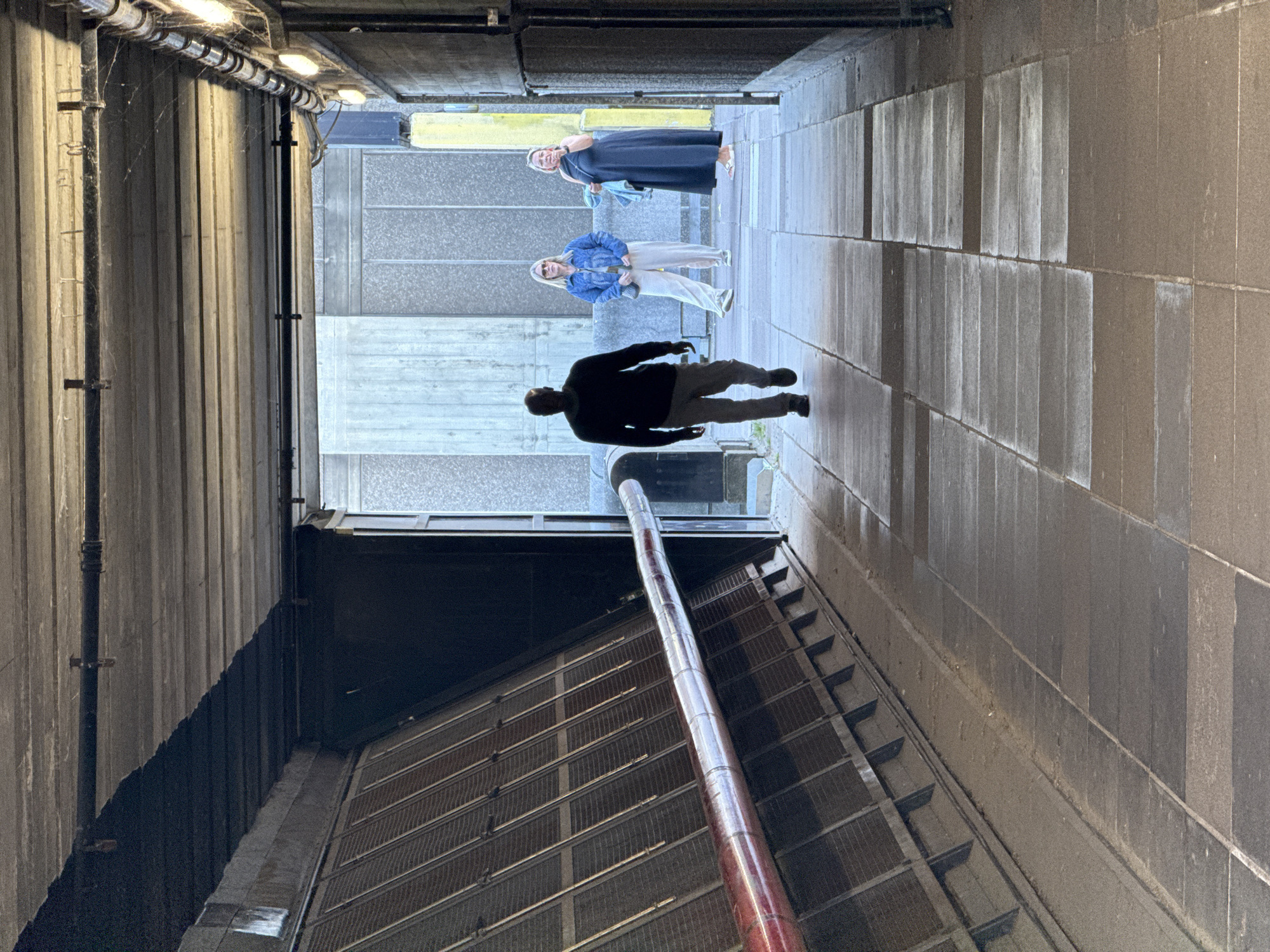 The image shows a pedestrian walkway, likely a short tunnel or covered passage, with three people walking in the distance. The person closest to the viewer is a male in dark clothing and light trousers. The second person is a female in a light-colored jacket and light trousers. The third person, slightly further away, is a female in a dark dress. The pathway is constructed of light gray stone blocks that form a narrow passage and the ceiling is lined with lights. One side of the walkway features a grid and dark overhang. The presence of lights and the style of the architecture suggest an urban setting in the United Kingdom, likely London.