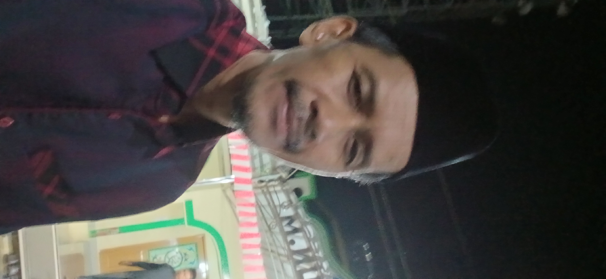 A man wearing a black peci cap and a red and black plaid shirt is looking at the camera. He has a mustache and a goatee. He is standing in front of a building with a green roof and white and pink striped awnings. In the background, another person can be seen walking away from the camera. The scene appears to be outdoors at night, possibly in Samarinda, Indonesia.