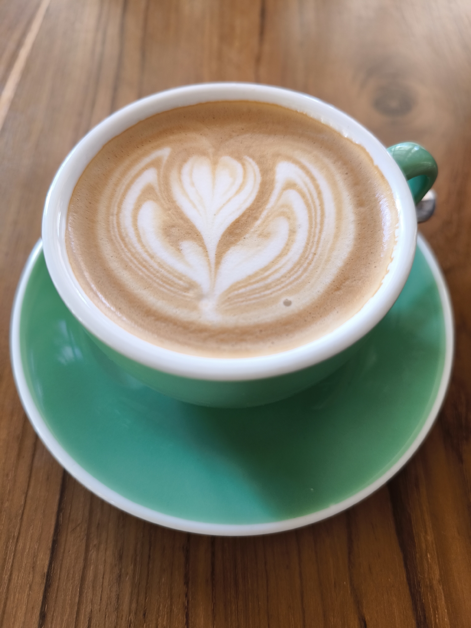 A high-angle, close-up shot showcases a cup of coffee with latte art in a green cup and saucer, sitting on a wooden table. The latte art features a heart-like pattern in the center. The setting is likely a cafe in Kuta, Indonesia. No other subjects are in the scene.