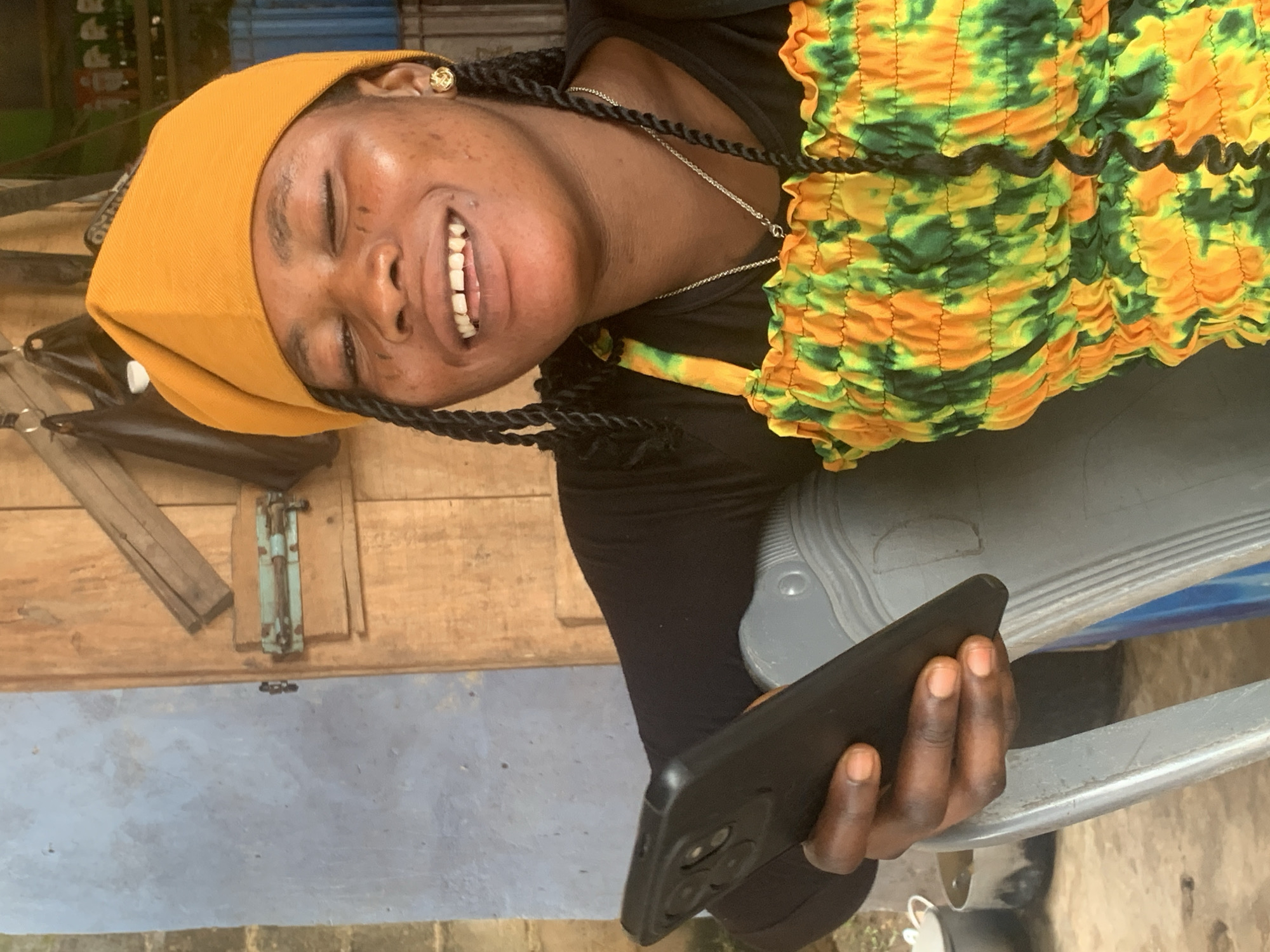 A young woman with her eyes closed and a wide smile is looking down at her black smartphone, which she holds in her left hand. She is wearing a black long-sleeved shirt and a ruched, sleeveless top with a tie-dye pattern of orange and green. A gold earring adorns her right ear, and a thin silver chain rests around her neck. She has black braided hair, with one braid visible over her right shoulder. She is seated on a grey chair in front of a wooden door. The background shows a textured blue wall to the left of the door and some shelves with bottles and bags on the right. The scene appears to be outdoors in a rustic setting, possibly in the city of Shagamu, Nigeria.