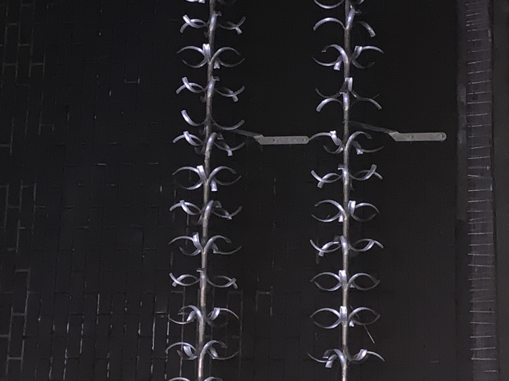 The image captures a dark, indoor scene featuring two prominent vertical metallic structures, likely decorative panels, fencing, or a screen. Each structure is adorned with numerous small, silvery, eye-shaped or loop-shaped metallic ornaments, arranged symmetrically along its length, creating a repetitive pattern. These polished elements reflect light, standing out against the somber background. A slender, flat, light-colored metallic bar connects the two vertical structures horizontally near their centers. The background is exceptionally dark and appears to be a textured wall, possibly brickwork or paneling, though its details are largely obscured by the low light. To the far right, a portion of another dark vertical element with distinct horizontal striations is visible. No human activity is depicted, and there is no visible text. The overall aesthetic is one of structured design or industrial artistry, with a focus on metallic patterns in a dimly lit environment, potentially an architectural detail within London.