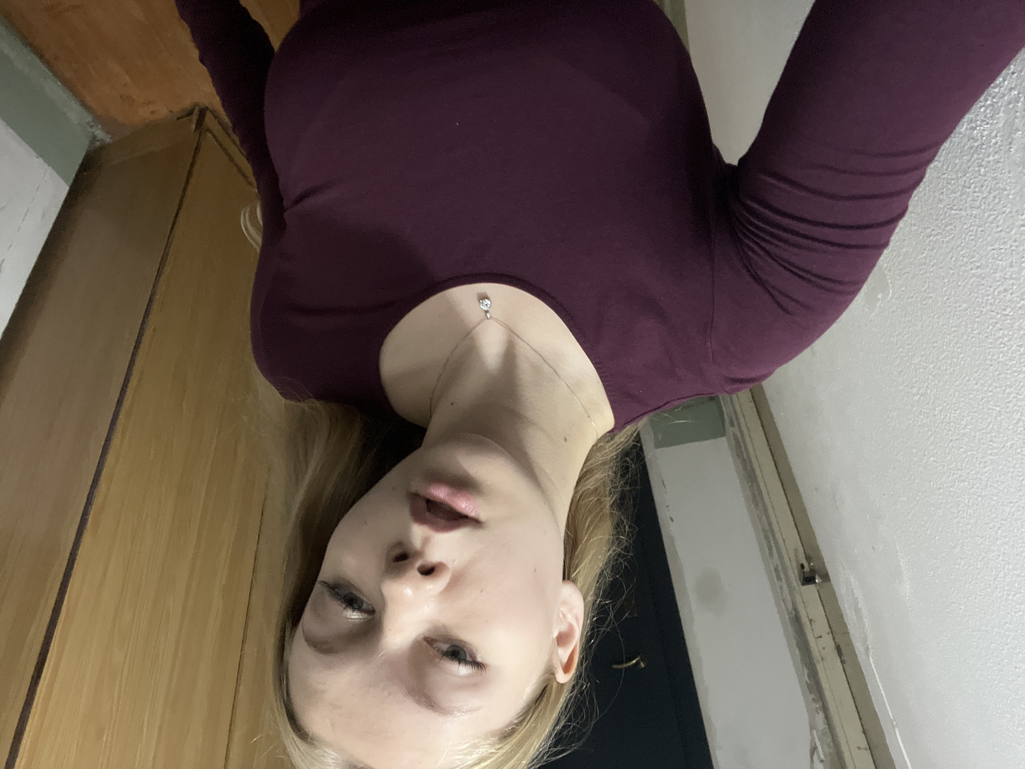 A young woman with blonde hair and fair skin is taking a selfie indoors. She is wearing a dark purple long-sleeved shirt and a simple silver necklace with a small pendant. Her face is turned slightly to the side, and she has light makeup on. The background suggests an indoor setting, possibly a hallway or a room with wooden doors and textured walls. The lighting is dim. The woman's pose and the overall composition of the image indicate a casual, personal photograph. The location is not explicitly visible in the image, so it cannot be determined to be in the city of Perm, Russia.