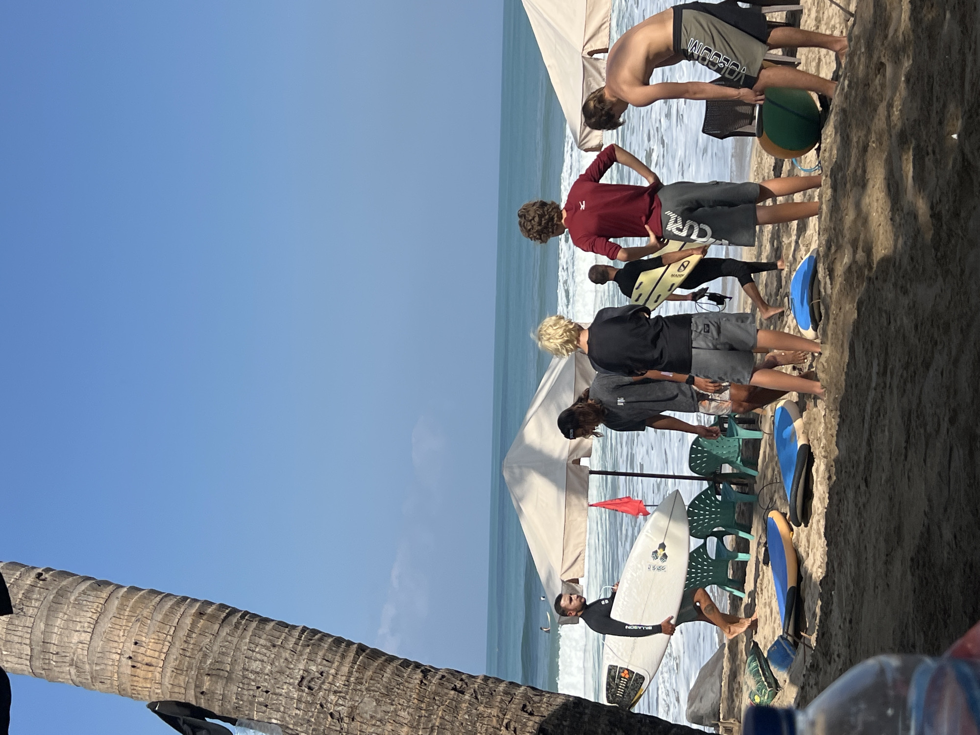 This is a medium-shot, eye-level, vertical photograph taken on a sunny day at the beach in Kuta, Indonesia. The foreground is dominated by sand with two surfboards lying on it, one blue and yellow, the other blue and white. In the middle ground, several young males are gathered near two large, off-white beach umbrellas. One man, wearing a black wetsuit, is carrying a white surfboard. Another young man in a black t-shirt and grey shorts is holding a yellow surfboard. A third person, wearing a maroon long-sleeved shirt and grey shorts, stands with hands on hips, facing away from the camera. Another young male, bare-chested and wearing patterned shorts, is facing the ocean. Two green plastic beach chairs are visible between the umbrellas. In the background, the turquoise ocean stretches to the horizon under a clear blue sky. Waves are breaking on the shore, and a few surfers can be seen in the water. A tall palm tree trunk is on the left side of the frame, casting a shadow.